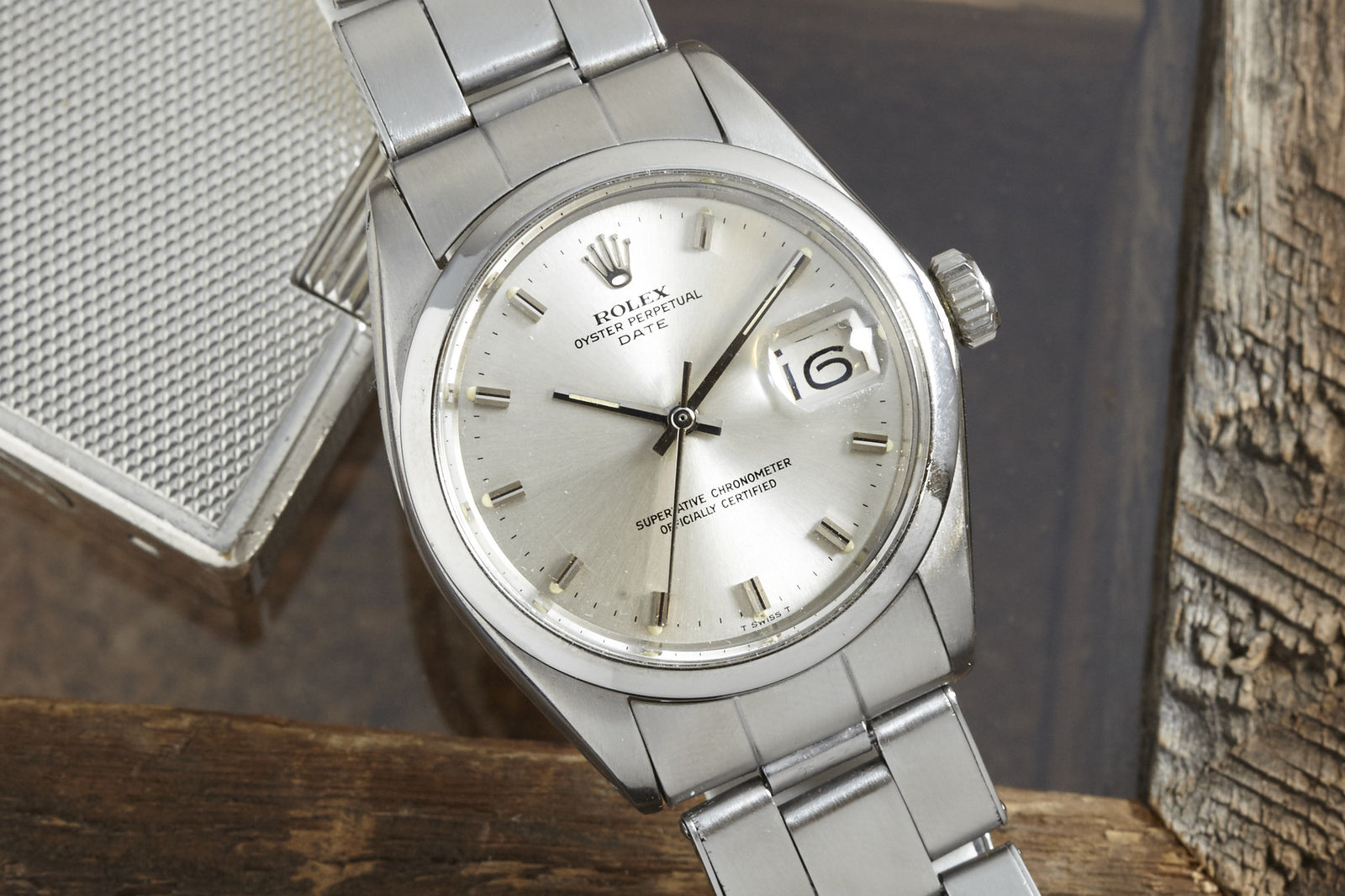 A Rolex Oyster Perpetual Date watch with a silver dial, steel rivet bracelet, and date window displaying "6" rests on a wooden surface near a metallic object.