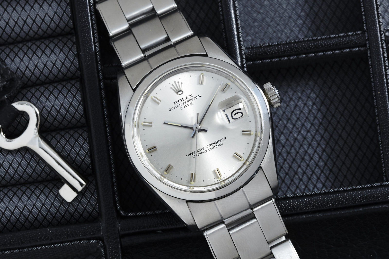 A silver Rolex Oyster Perpetual Date by Rolex with a steel bracelet is displayed on a black textured surface beside a metal key, showing the date as 16 and featuring minimalist hour markers.