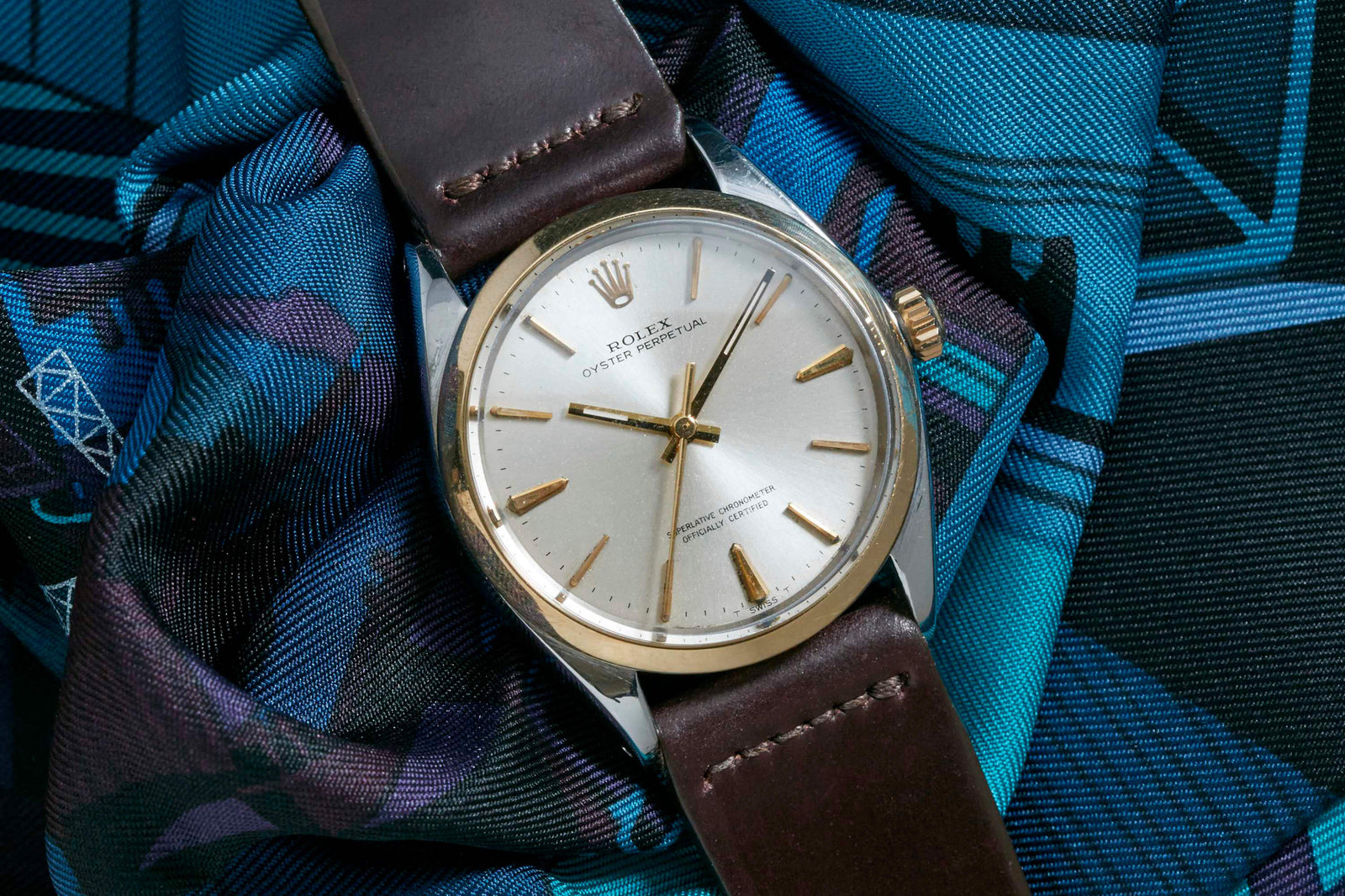 The Rolex Oyster Perpetual watch by Rolex, featuring a silver dial, gold markers, and brown leather strap, is displayed on blue patterned fabric, highlighting its classic Rolex Oyster case design.