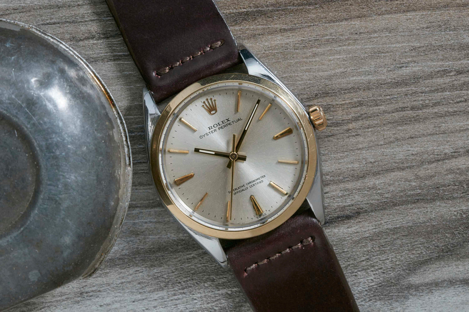 A close-up of the Rolex Oyster Perpetual by Rolex, featuring its signature Oyster case, gold bezel, silver dial, and brown leather strap, displayed on a wooden surface beside a metallic object.