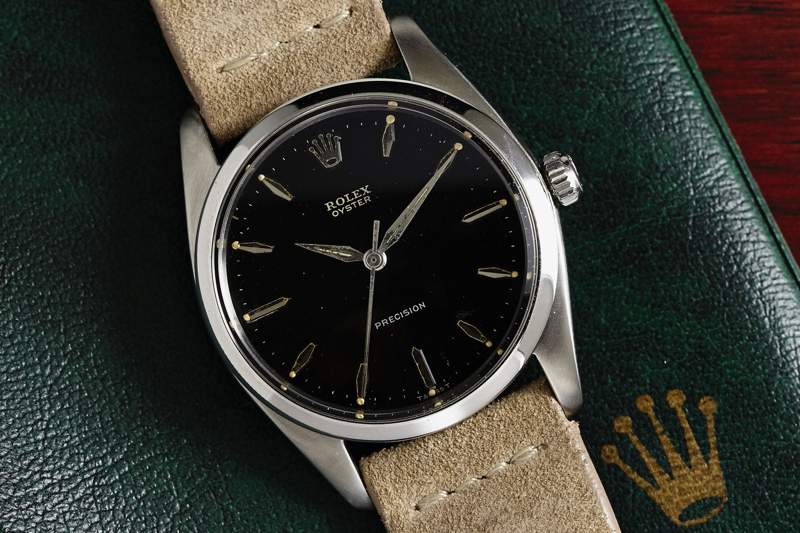 The Rolex Oyster Precision - 1959 by Rolex, featuring a black dial, silver hands, and a beige suede strap, rests on green leather adorned with the gold Rolex crown logo.