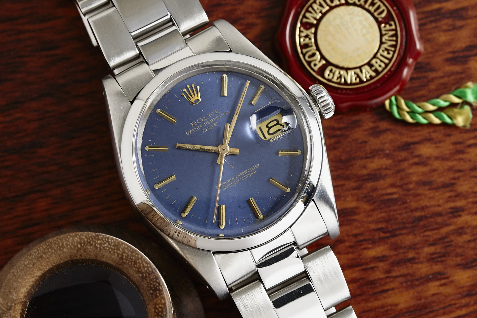 A close-up of a 1974 Rolex Oyster Perpetual Date by Rolex, featuring a blue sunburst dial and date display, rests on a wooden surface beside a wax seal and green-gold string.