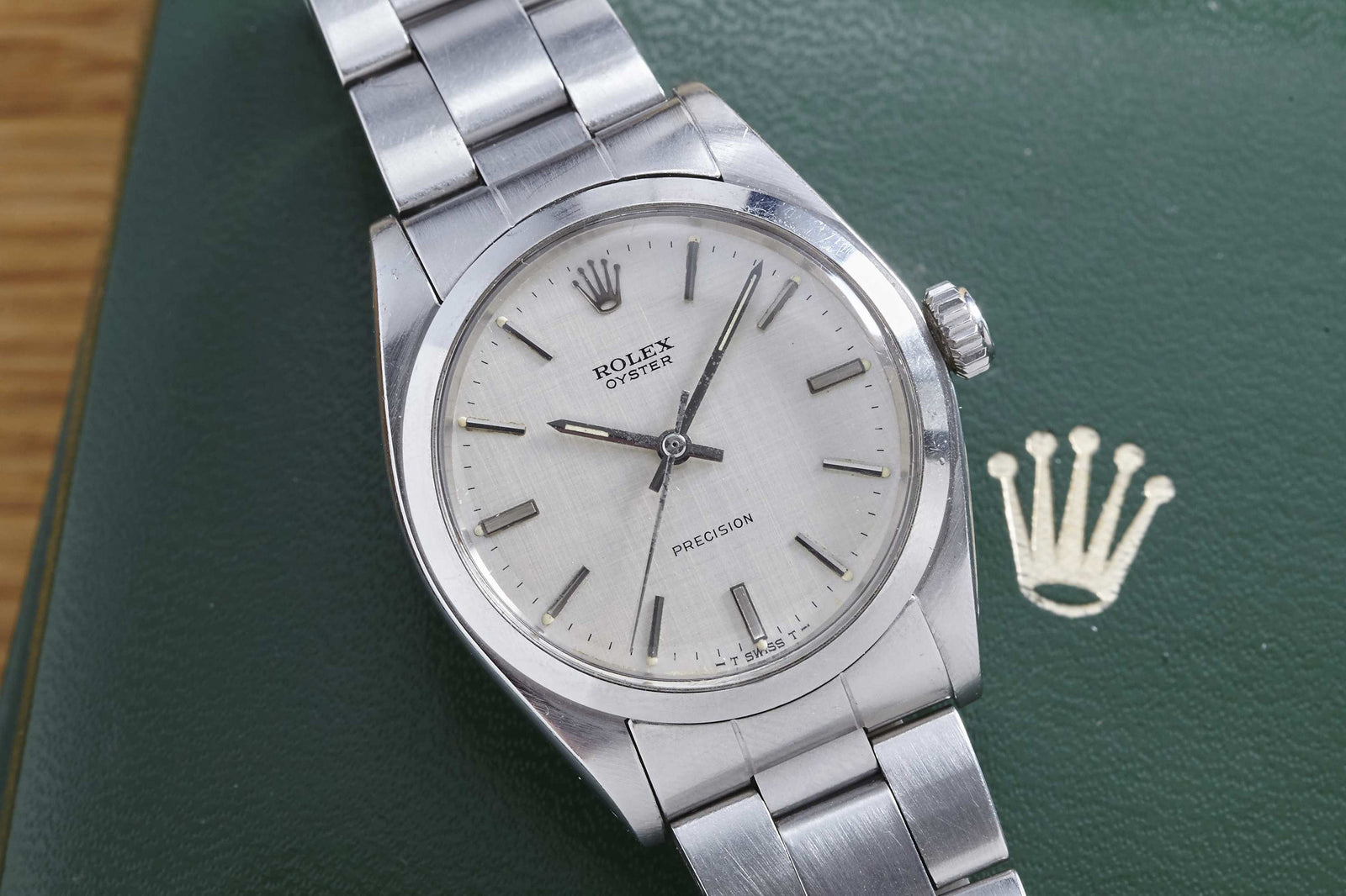 A Rolex Oyster Precision Linen Dial wristwatch, featuring a silver case and metal band, sits atop a green box with a gold Rolex crown logo.