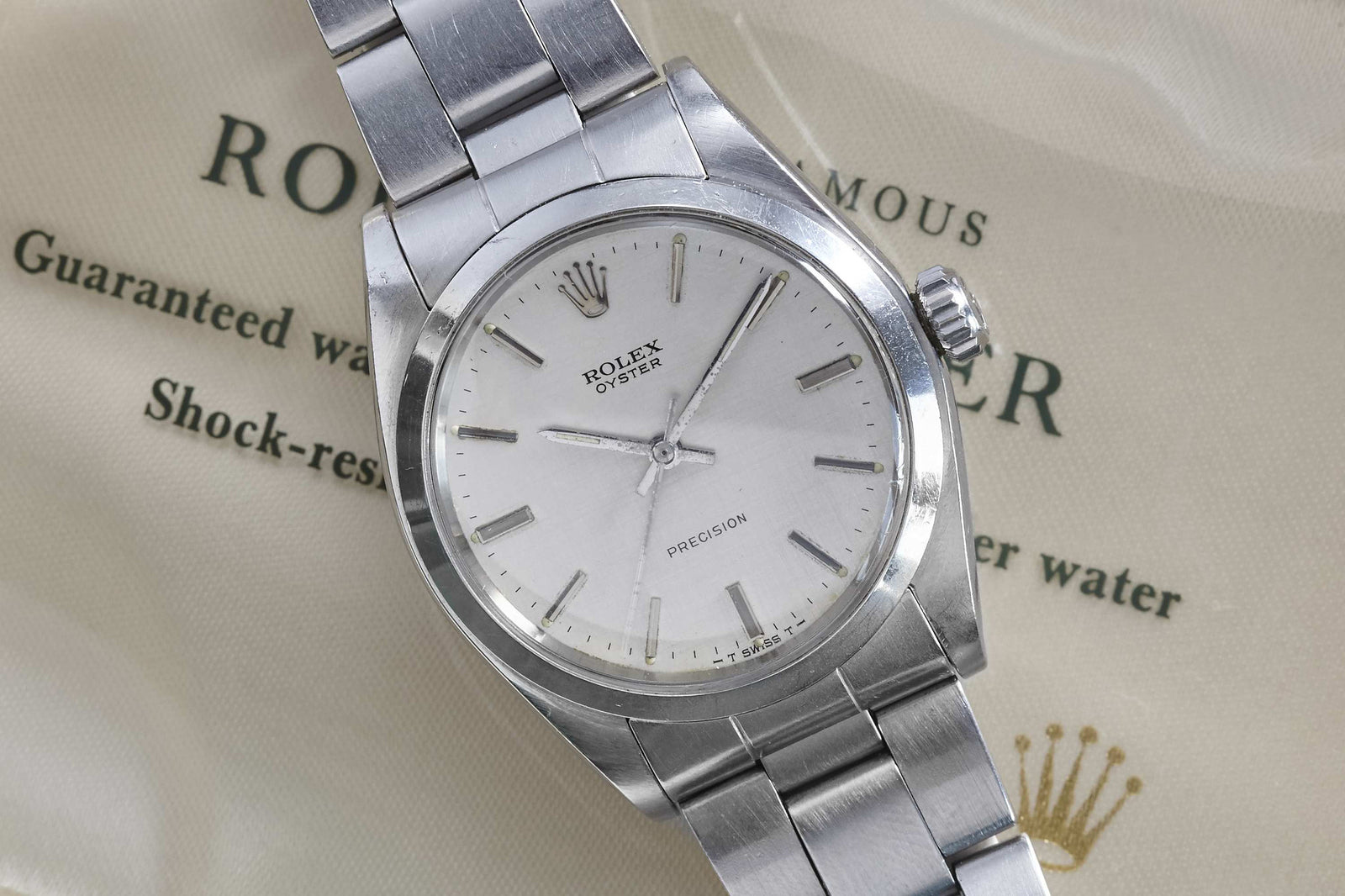A Rolex Oyster Precision Linen Dial wristwatch with a metal band rests on a cream Rolex document with green text and a gold crown logo, showcasing the refined style of this classic timepiece from Rolex.