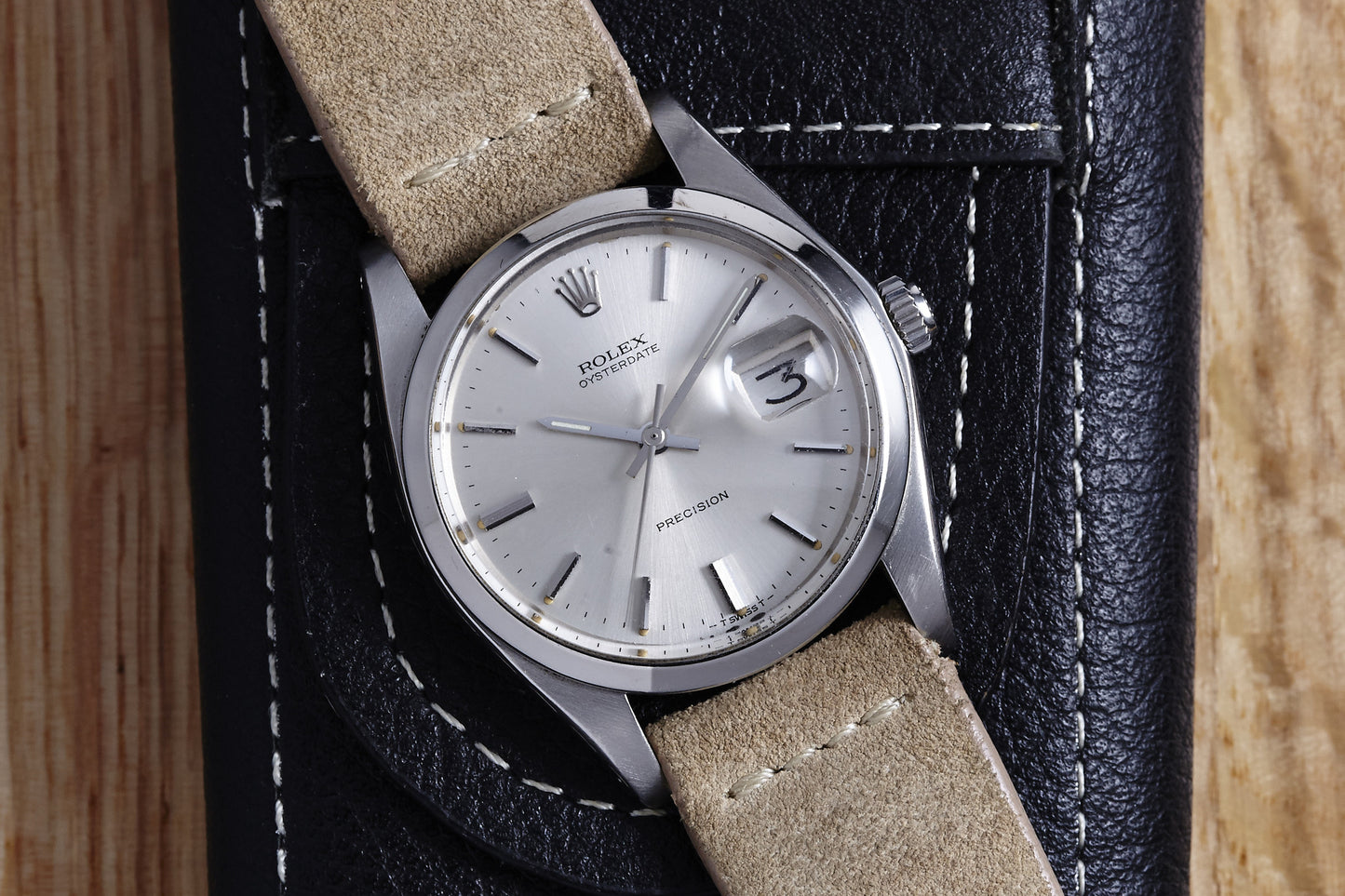 A 1978 Rolex Oysterdate Precision silver Swiss watch with a beige leather strap and date display rests on a black leather case on a wooden surface.