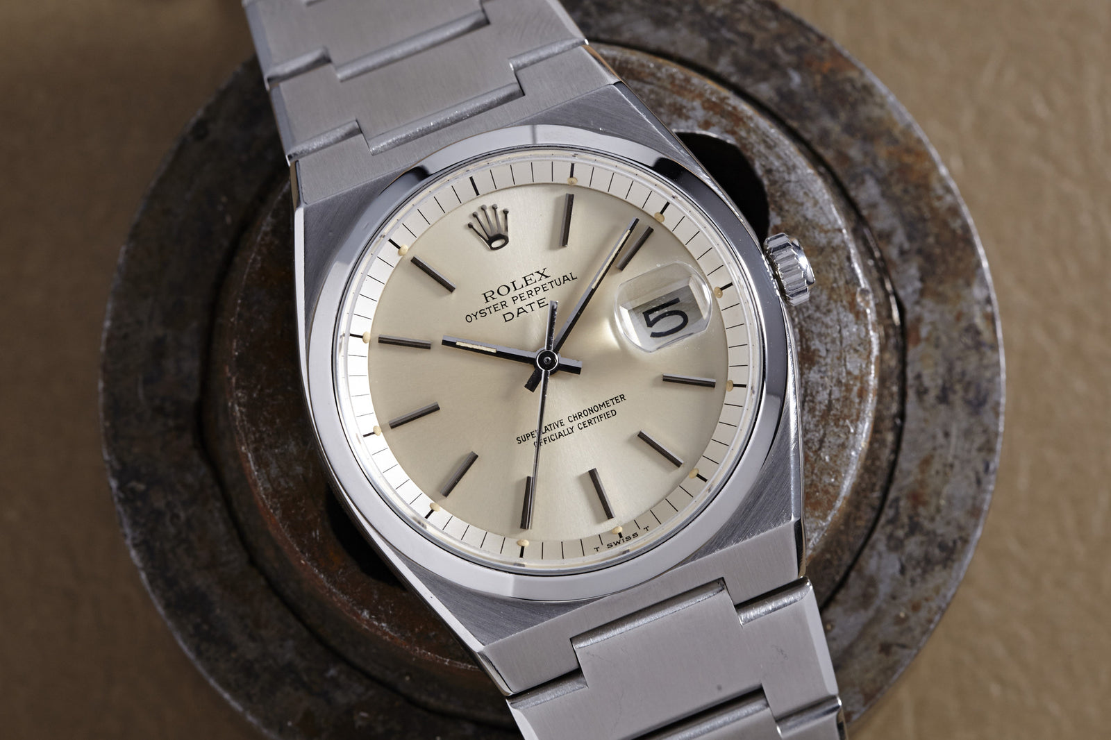 The Rolex Reference 1530 Oyster Perpetual Date by Rolex, featuring a cream dial and date window at 3 o’clock, rests elegantly on a round, weathered metallic surface.