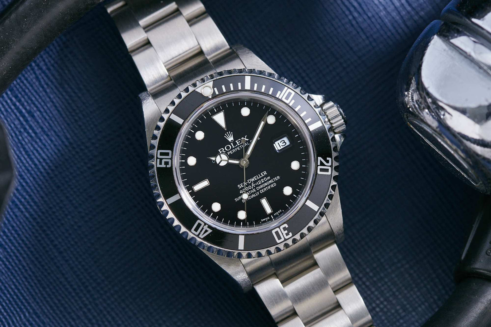 A Rolex Sea-Dweller professional diver’s watch with a black dial and bezel is displayed on a dark textured surface beside metallic and black objects. The time reads 10:10 with the date set to the 13th.
