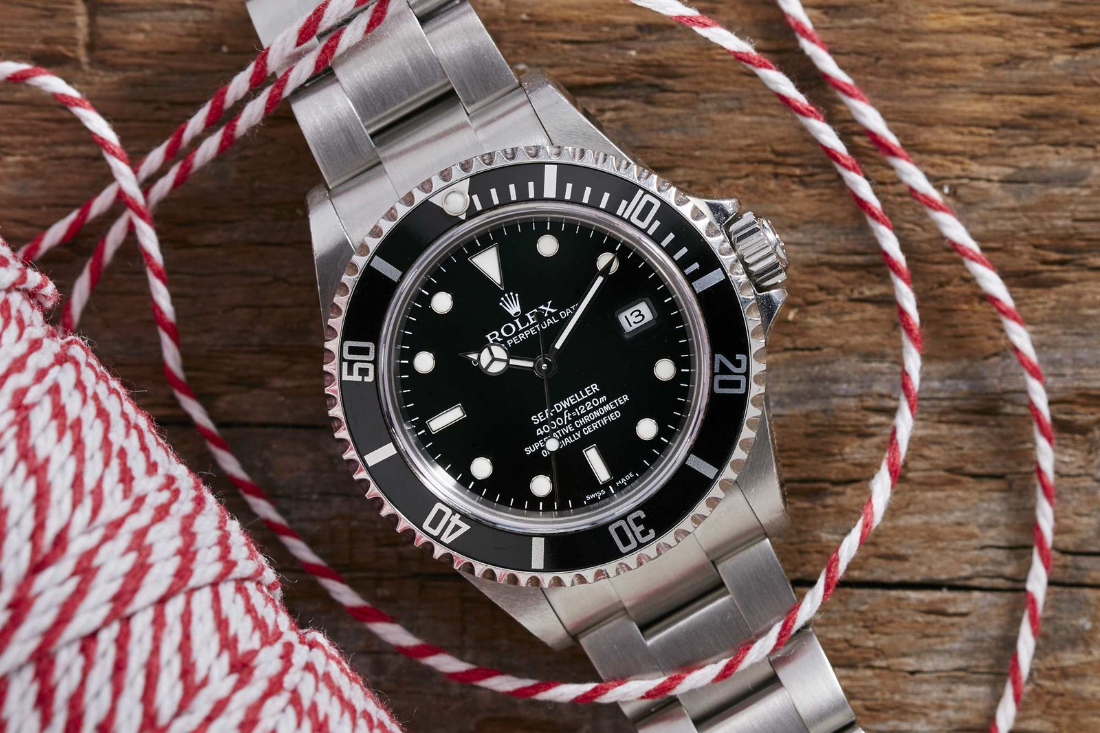 The Rolex Sea-Dweller, a stainless steel professional diver's watch with black dial and bezel, rests on a wooden surface surrounded by red and white striped twine. The time on this Rolex masterpiece reads 10:10.