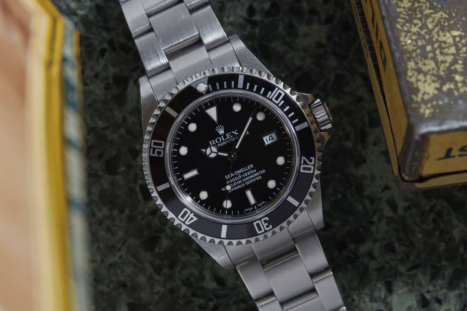 A close-up of the Rolex Sea Dweller by Rolex, a stainless steel watch with black dial and date window, designed for saturation diving and used by COMEX divers, placed on a green marbled surface next to a yellow box.