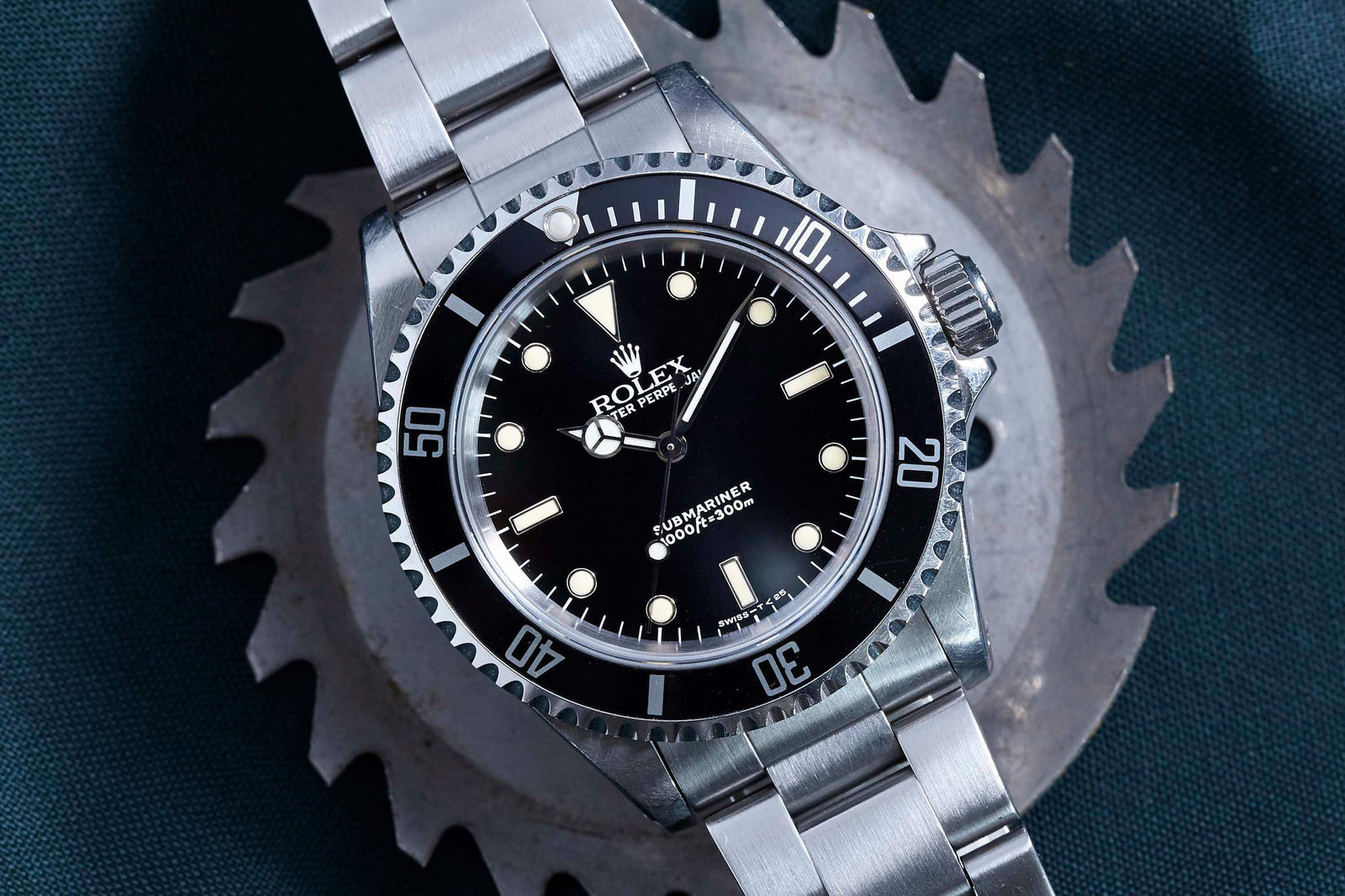 The Rolex Submariner by Rolex, crafted in stainless steel with a black dial and bezel, is showcased on a metallic gear against a dark green backdrop.