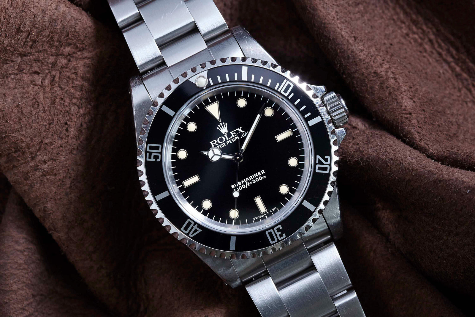 A close-up of the Rolex Submariner by Rolex, featuring a black dial, white hour markers, and a stainless steel bracelet, displayed on brown textured fabric.