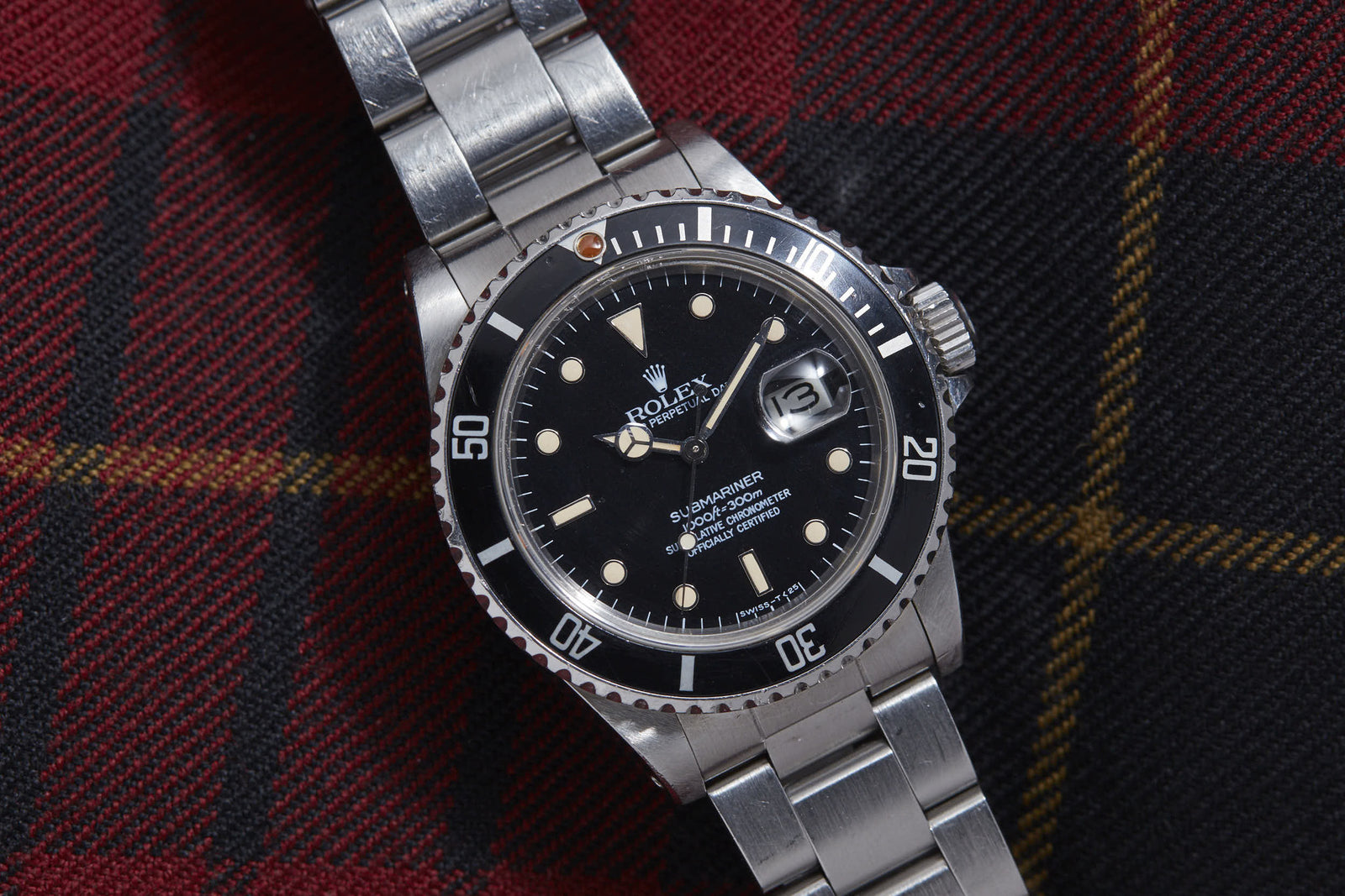 The Rolex Submariner Reference 16800 by Rolex, featuring stainless steel, a matte black dial and bezel, and a date display, sits on dark plaid fabric—an ideal pick for vintage watch enthusiasts.