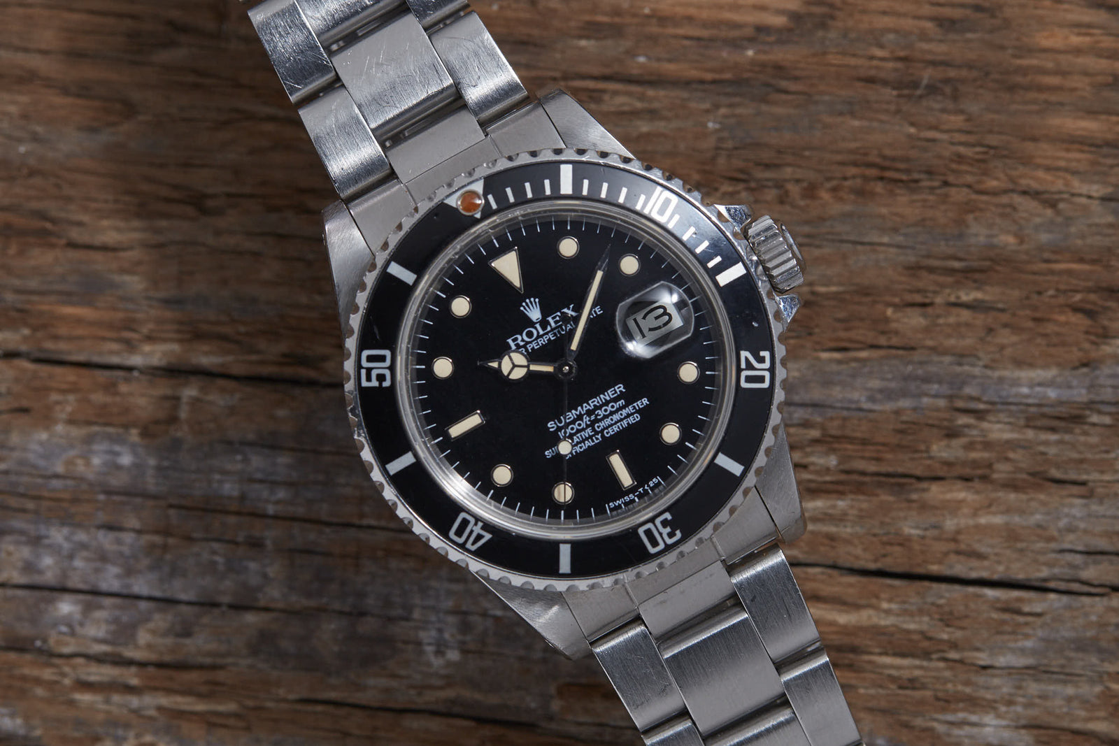 The Rolex Submariner Reference 16800 by Rolex features a stainless steel case, matte black dial with silver hour markers, and a metal bracelet, elegantly displayed on rustic wood.