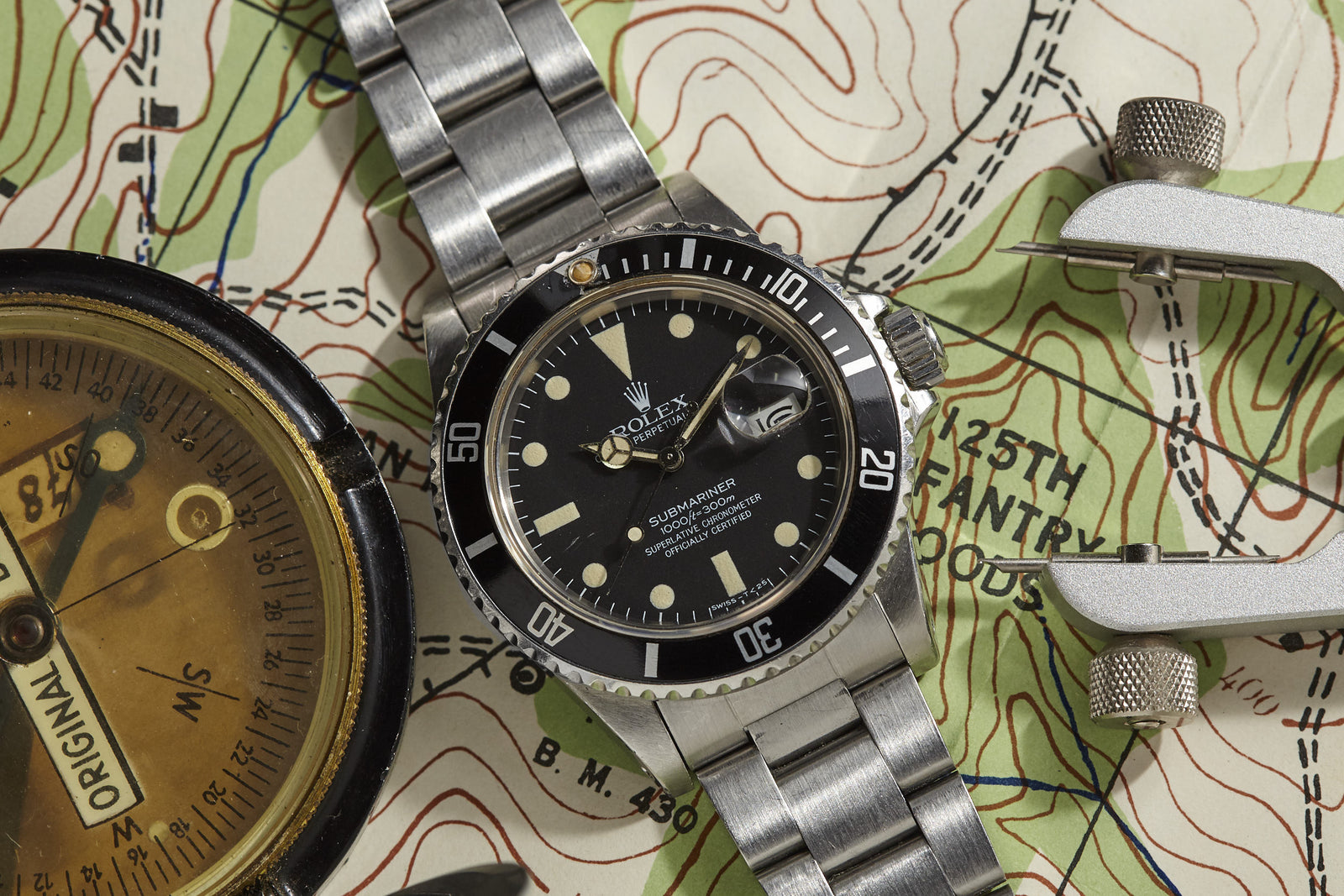 A Rolex Submariner by Rolex rests on a topographic map next to a brass compass and caliper, evoking a sense of navigation and adventure.