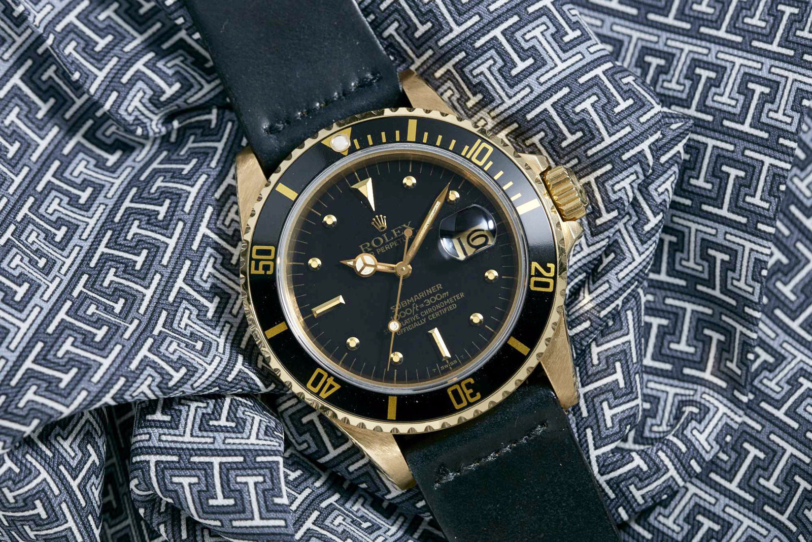 A Rolex Submariner by Rolex featuring a black bezel and black leather strap is displayed on patterned black and white fabric.