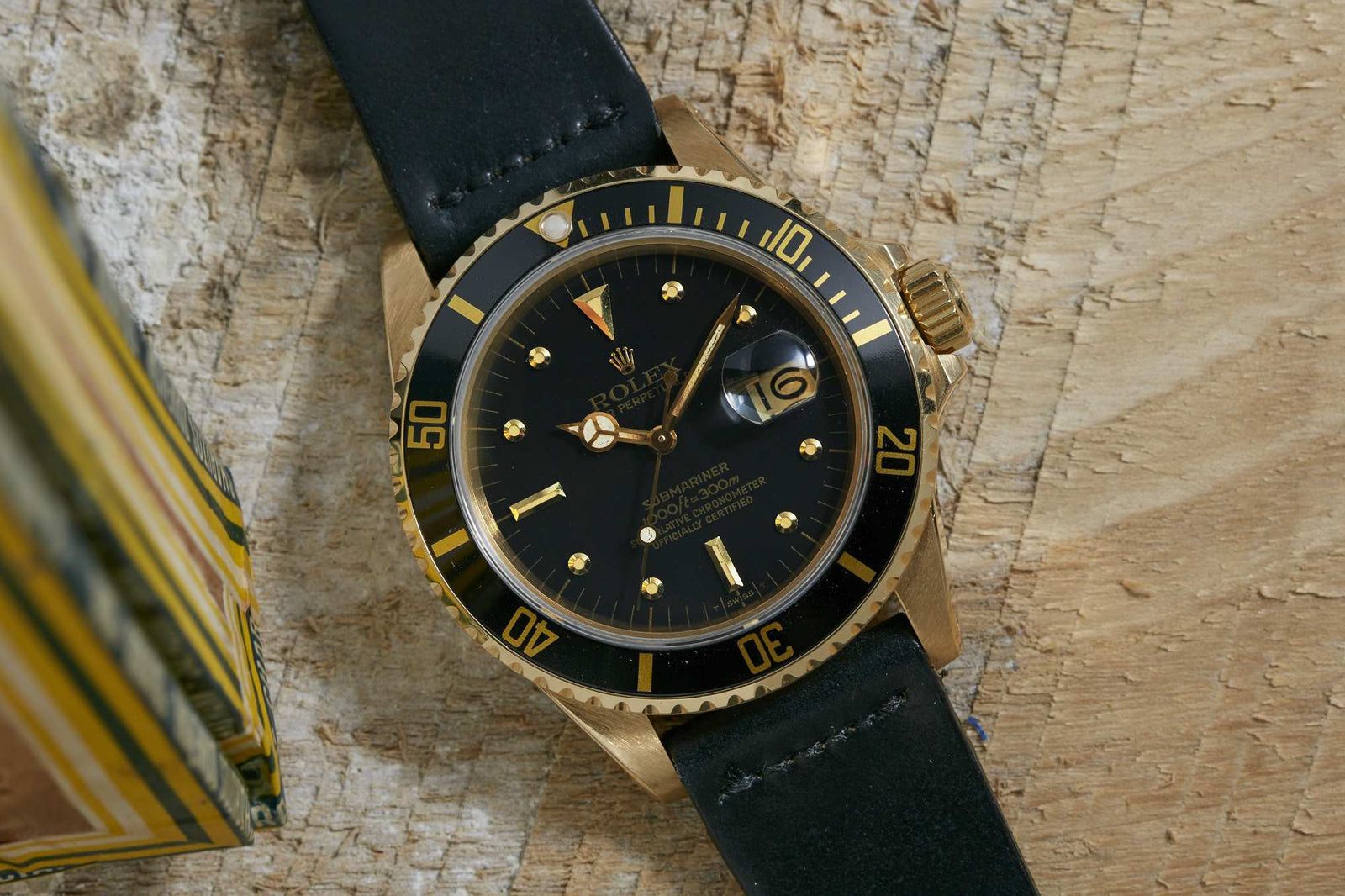 A solid gold Rolex Submariner by Rolex, featuring a black dial and bezel with gold hour markers and hands, is displayed on a textured wooden surface next to a striped yellow box.