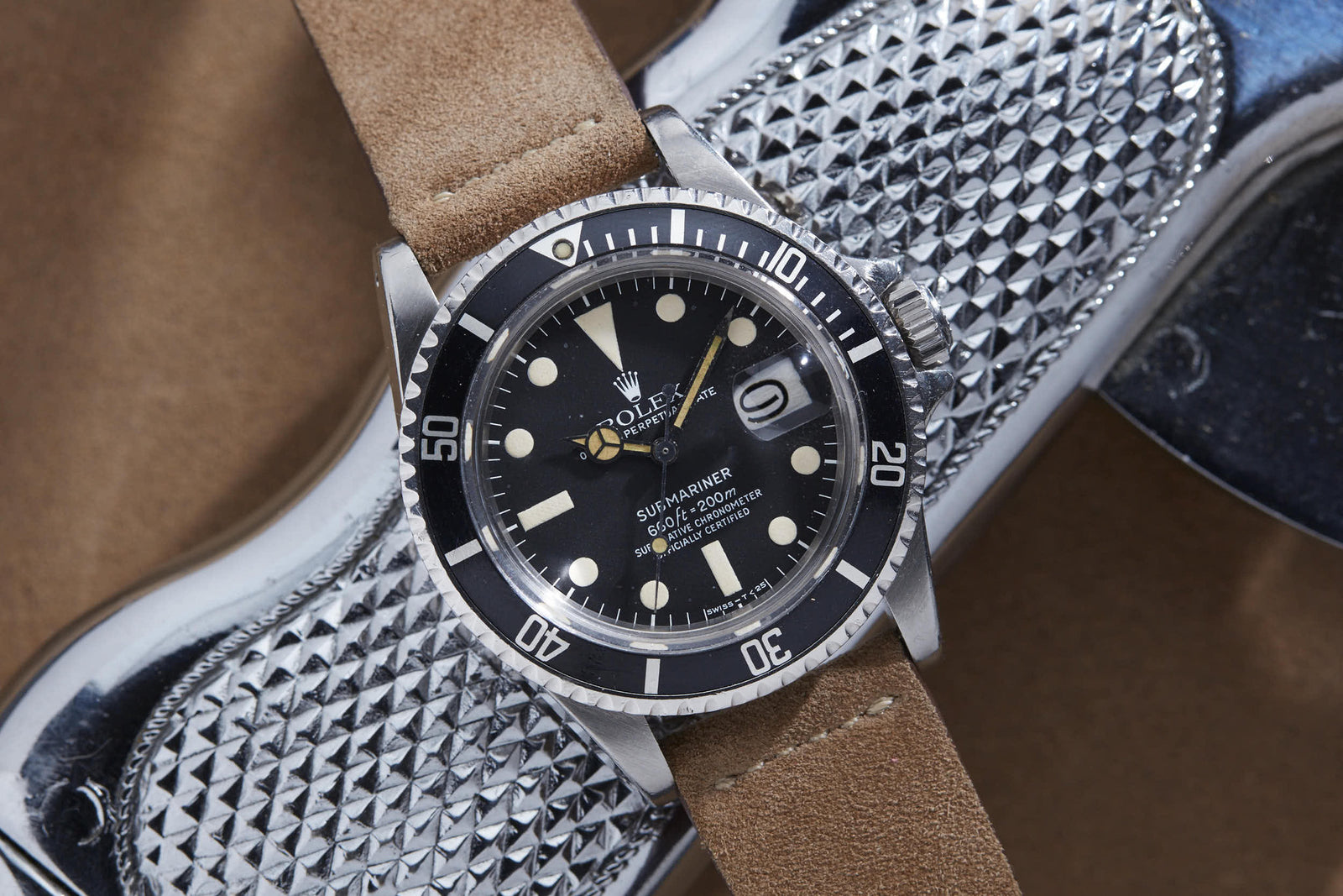 A Rolex Submariner watch with a black dial and tan leather strap from Rolex is displayed on a textured metallic surface.