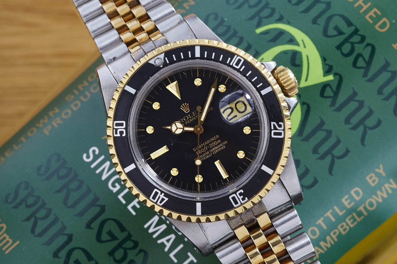 A close-up of the Rolex Submariner 1680 Two Tone Nipple Dial, featuring a black dial, two-tone bracelet, and magnified date window at 20, resting on a green box labeled Springbank Single Malt.