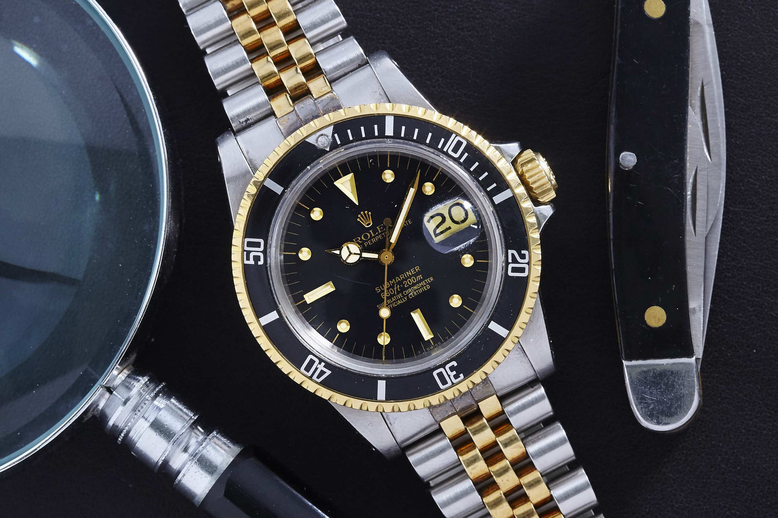 A close-up of a Rolex Submariner 1680 Two Tone Nipple Dial by Rolex, featuring a black dial with gold accents, resting on a dark surface beside a magnifying glass and a pocket knife.