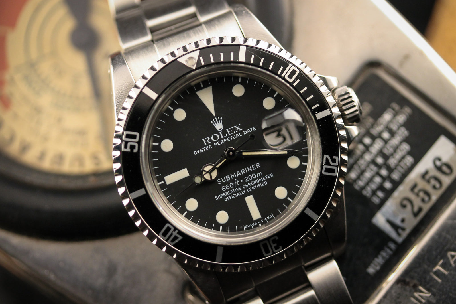 A close-up of a vintage Rolex Submariner 1680 -1979 wristwatch with a stainless steel band, black bezel, and dial rests on a surface, with a gauge and metal plate marked A-2556 in the background.