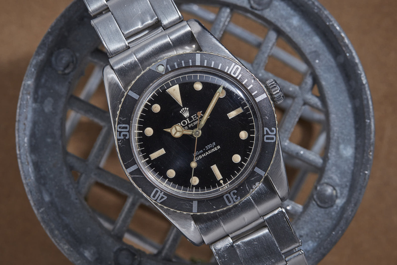 A Rolex Submariner sports watch by Rolex with a black dial, silver metal band, iconic logo, and luminous hour markers is displayed on a metal grate background.