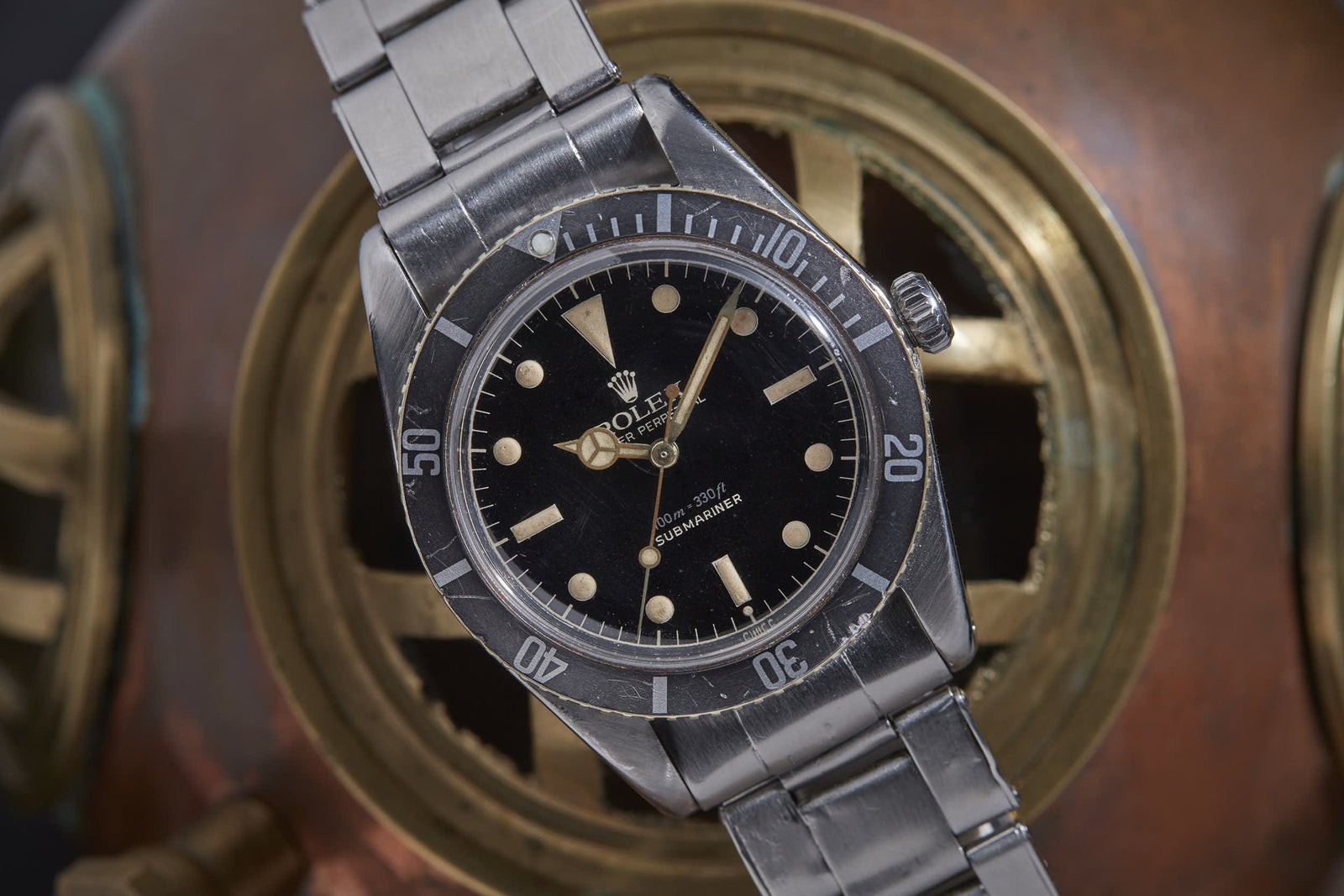 The Rolex Submariner by Rolex is a vintage sports watch featuring a black dial and metal bracelet, showcased against a brass, nautical-inspired background.