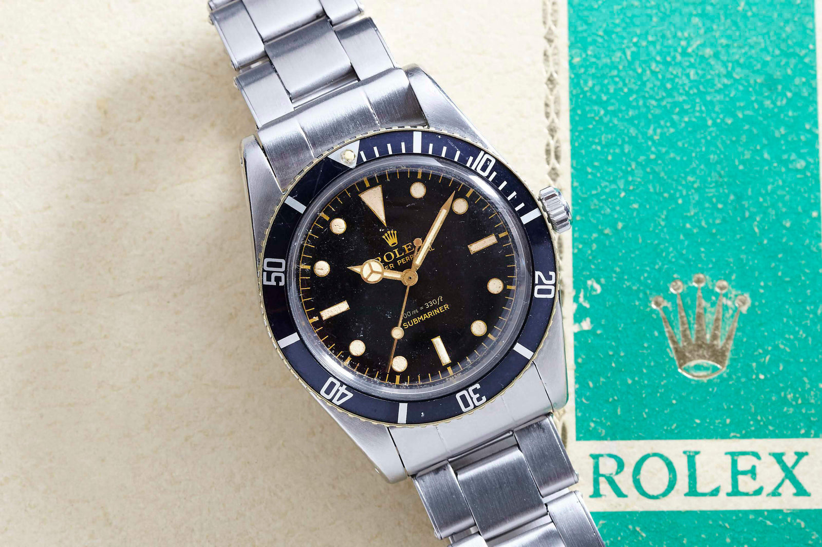 A vintage Rolex Submariner 5508 dive watch with a black bezel and dial rests on a light surface beside a worn green Rolex booklet featuring the gold crown logo and "ROLEX.