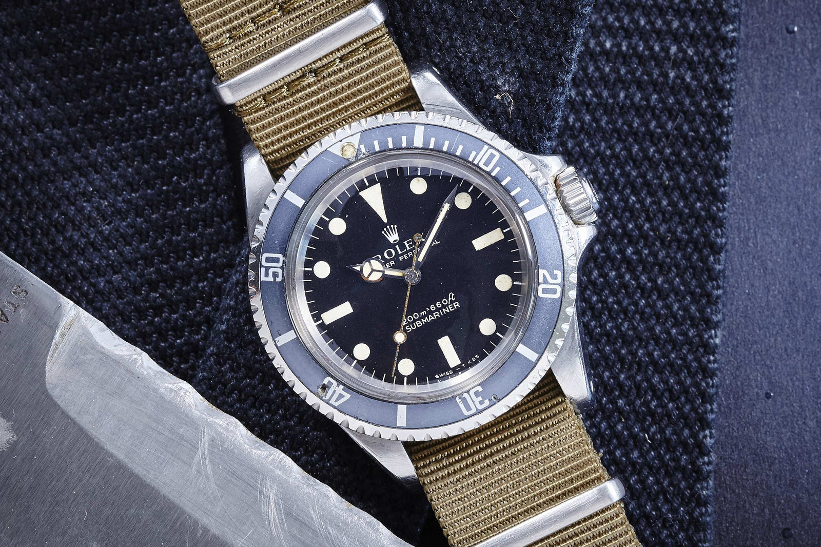 A Rolex Submariner wristwatch by Rolex, featuring a black dial, round indices, and a khaki NATO strap, rests on textured dark fabric beside a metallic object—an iconic tool watch with enduring style.