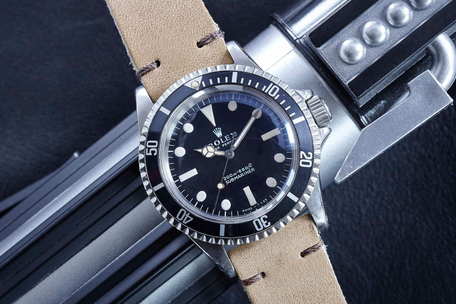 A Rolex Submariner by Rolex with a beige leather strap is displayed on a metallic, sci-fi-inspired object featuring buttons and grooves, set against a dark background.
