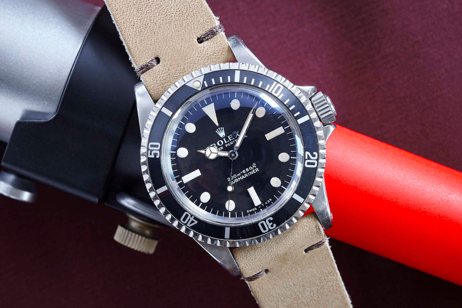 A Rolex Submariner by Rolex with a beige leather strap is displayed atop a black and red cylindrical object, set against a dark burgundy background.