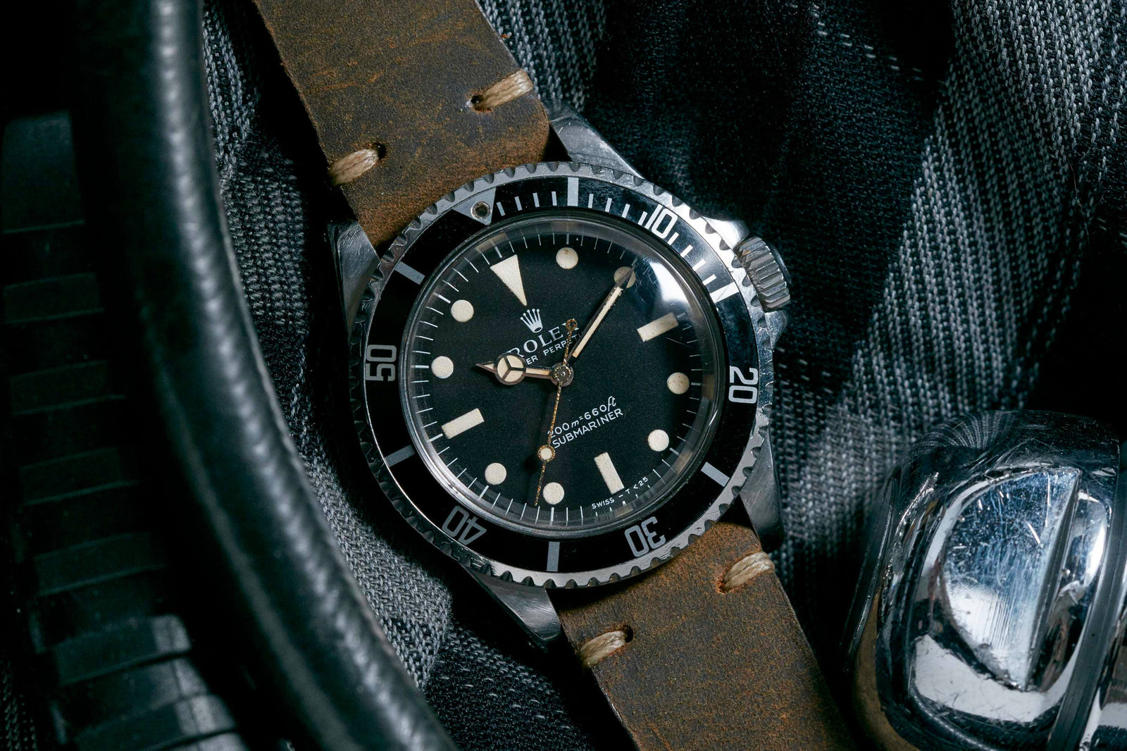 A Rolex Submariner by Rolex with a black dial, round hour markers, and a brown leather strap is displayed on dark textured fabric beside a metallic item.