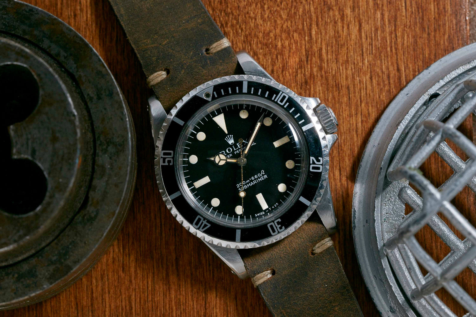 A Rolex Submariner by Rolex, featuring a black dial and bezel with a brown leather strap, is displayed on wood next to metallic mechanical parts.