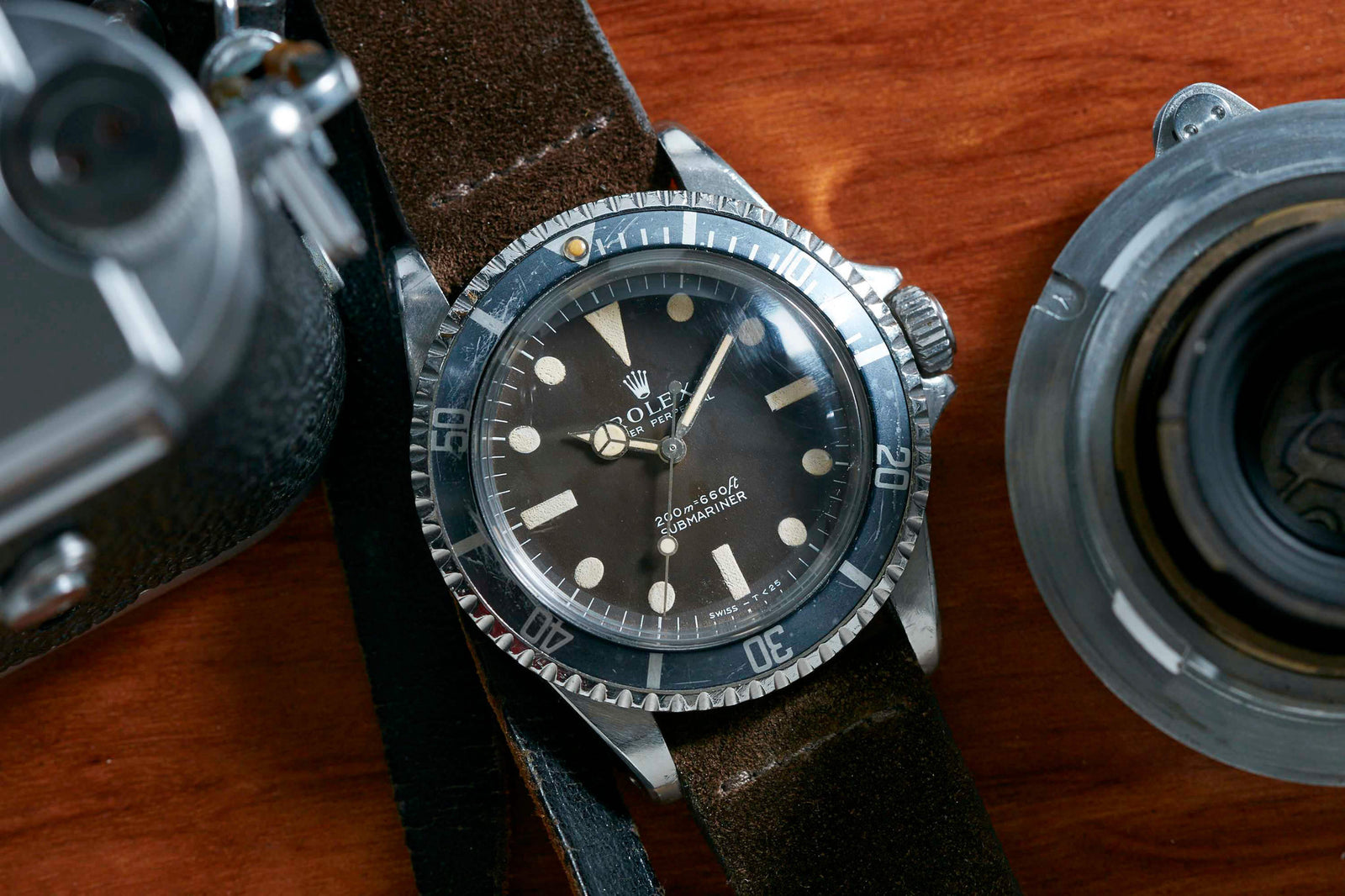 A Rolex Submariner Tropical Dial, featuring a black dial, black bezel, white markers, and silver hands on a brown leather strap with an Oyster case, rests on wood amid vintage camera parts.
