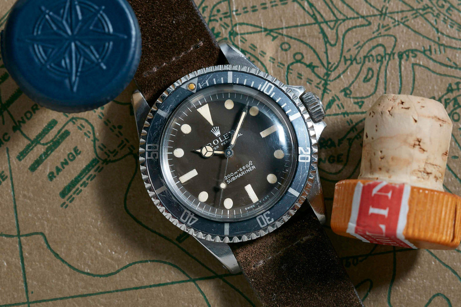 A Rolex Submariner Tropical Dial by Rolex with an Oyster case, black bezel, and brown leather strap rests on a map next to a blue wax seal stamp and a cork featuring a red and white stamp.