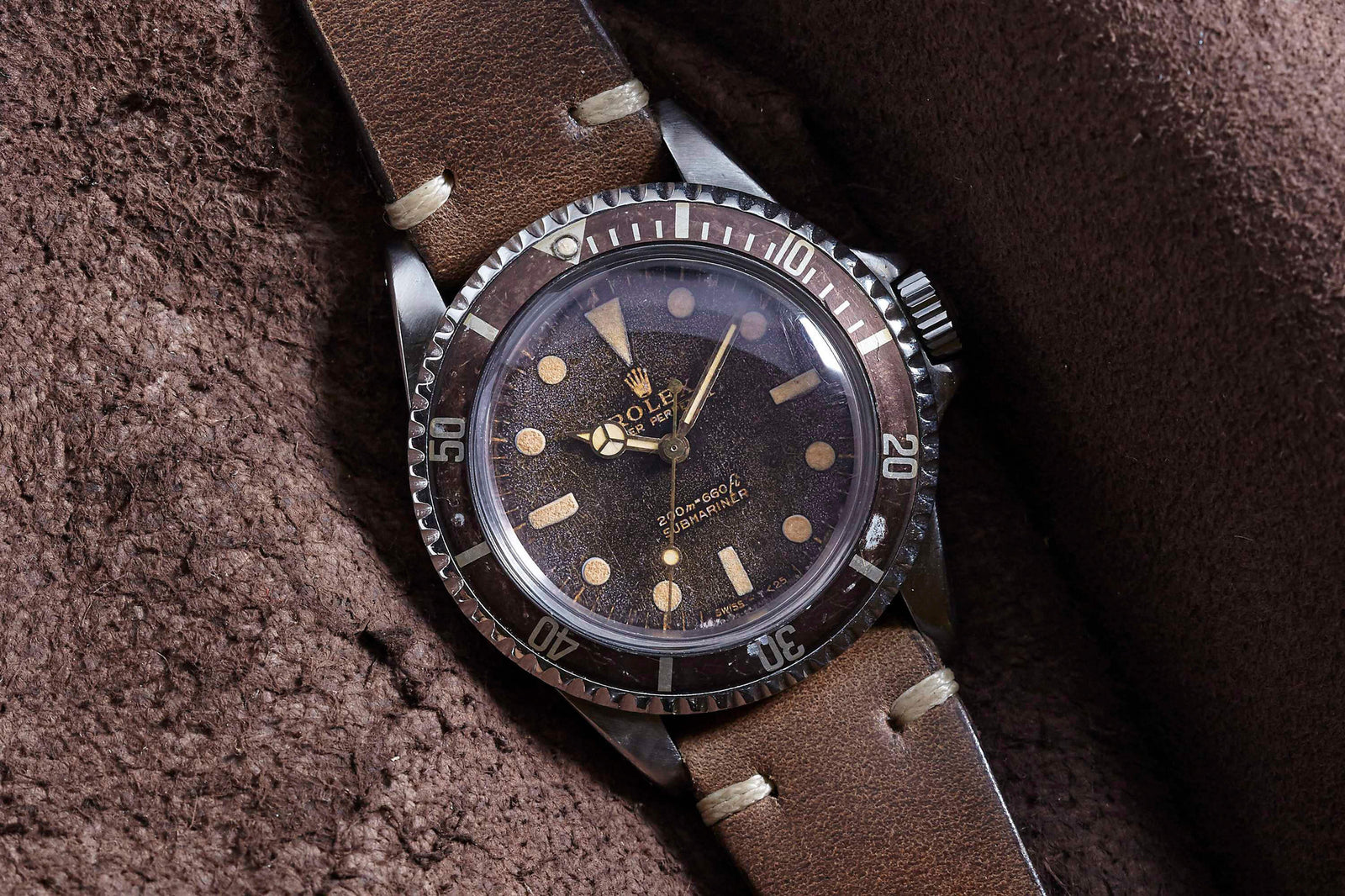 The Rolex Submariner by Rolex, with a brown dial, patina, large circular hour markers, and a brown leather strap, sits elegantly on textured brown fabric—a classic choice for watch enthusiasts.