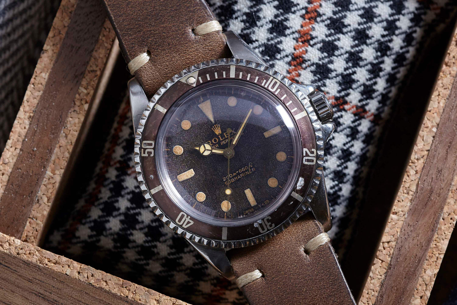 A Rolex Submariner by Rolex with a brown leather strap and worn dark bezel rests in a wooden box. It features beige hour markers and hands, with a houndstooth-patterned fabric in the background.