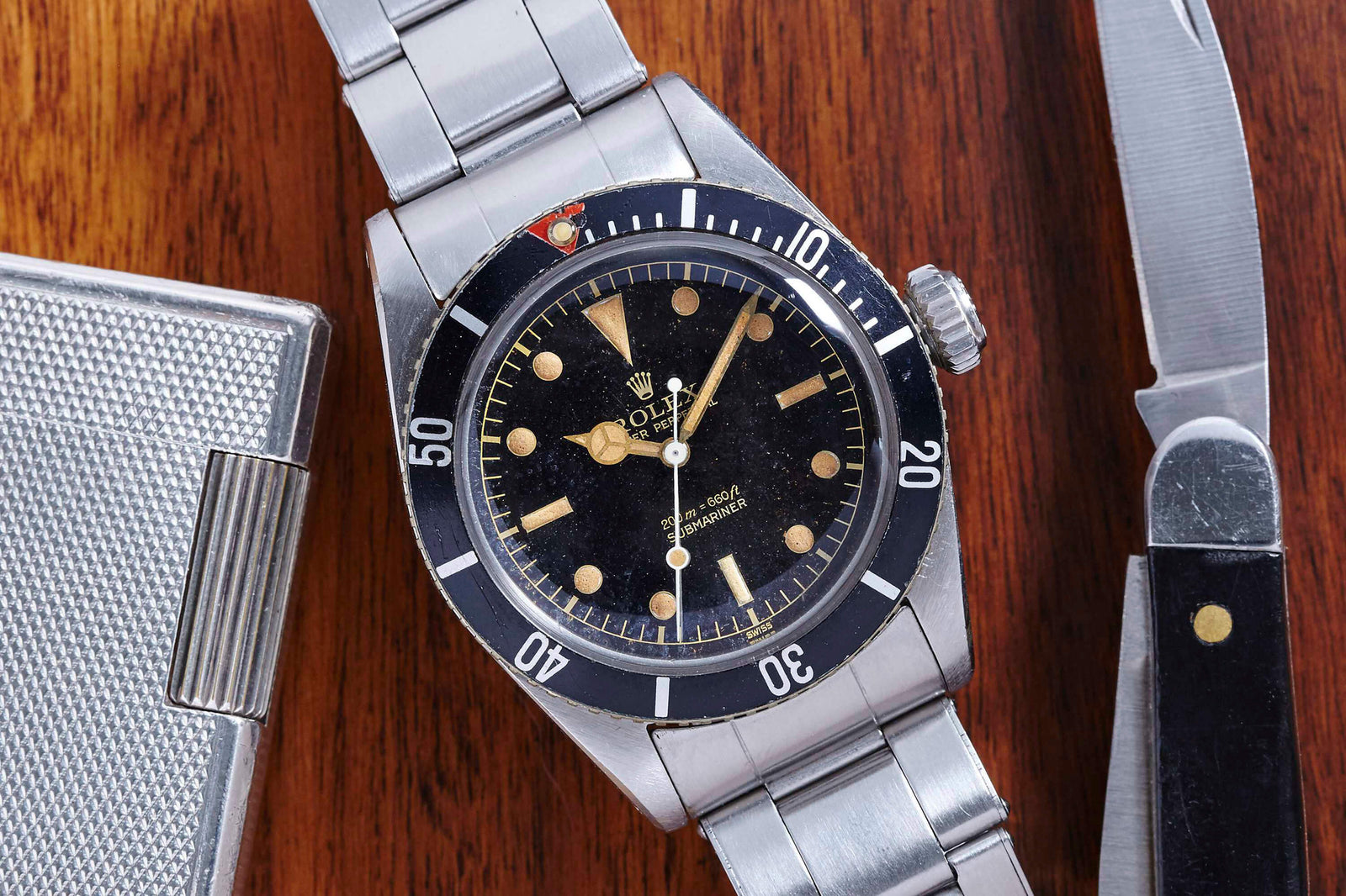 A Rolex Submariner 5510 "James Bond" by Rolex, featuring a black bezel and dial, is displayed on a wooden surface beside a metal lighter and folding knife. Detailed description coming soon!.