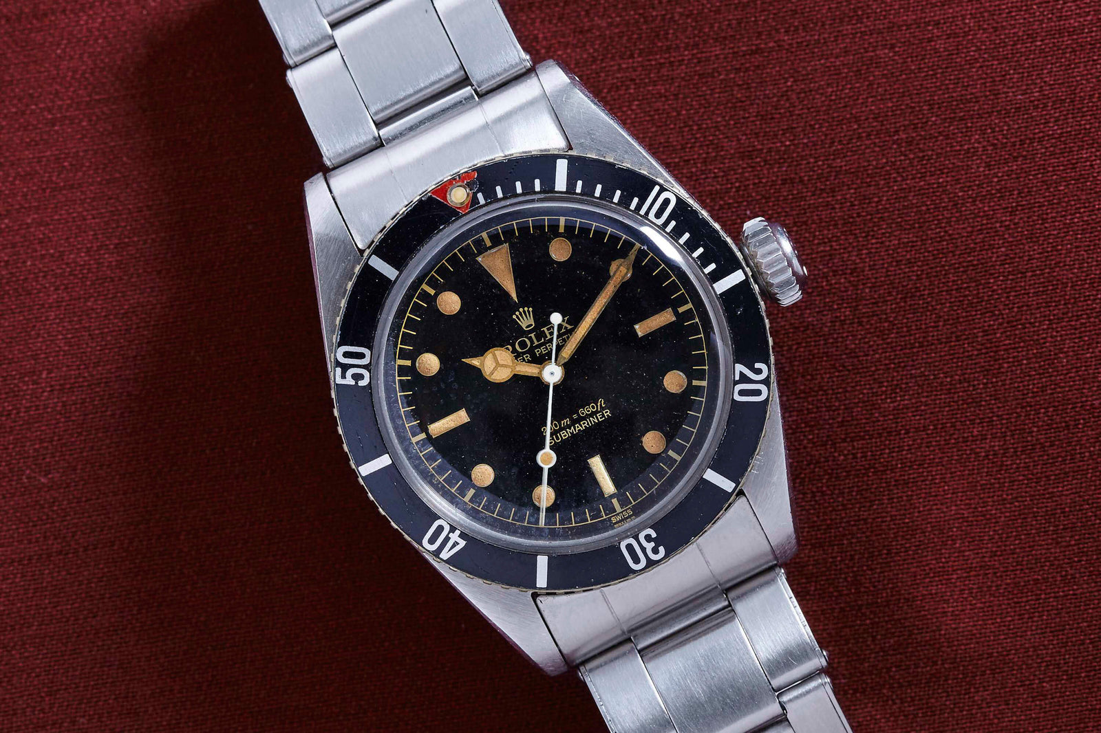A Rolex Submariner 5510 "James Bond" wristwatch with a black dial, round hour markers, and a stainless steel band is displayed on textured burgundy fabric. Detailed description coming soon!.