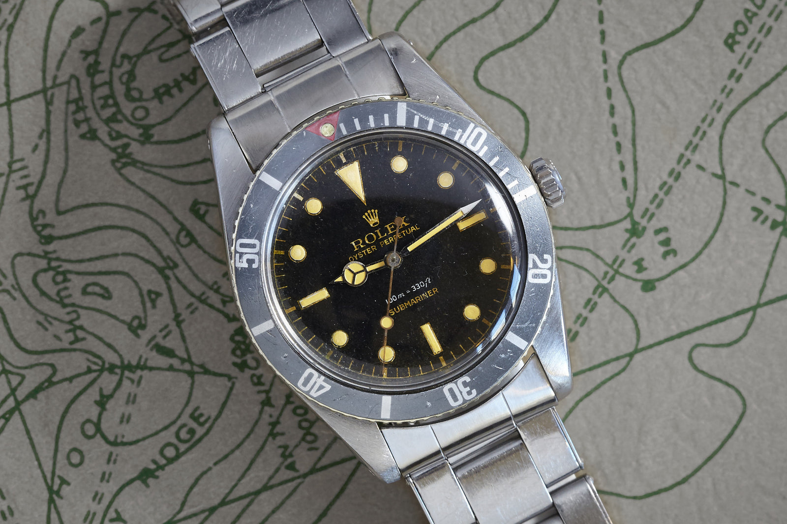 The Rolex Submariner Gilt Dial by Rolex features a black gilt dial, luminous markers, and a stainless steel bracelet, elegantly presented against a green and gray map backdrop.