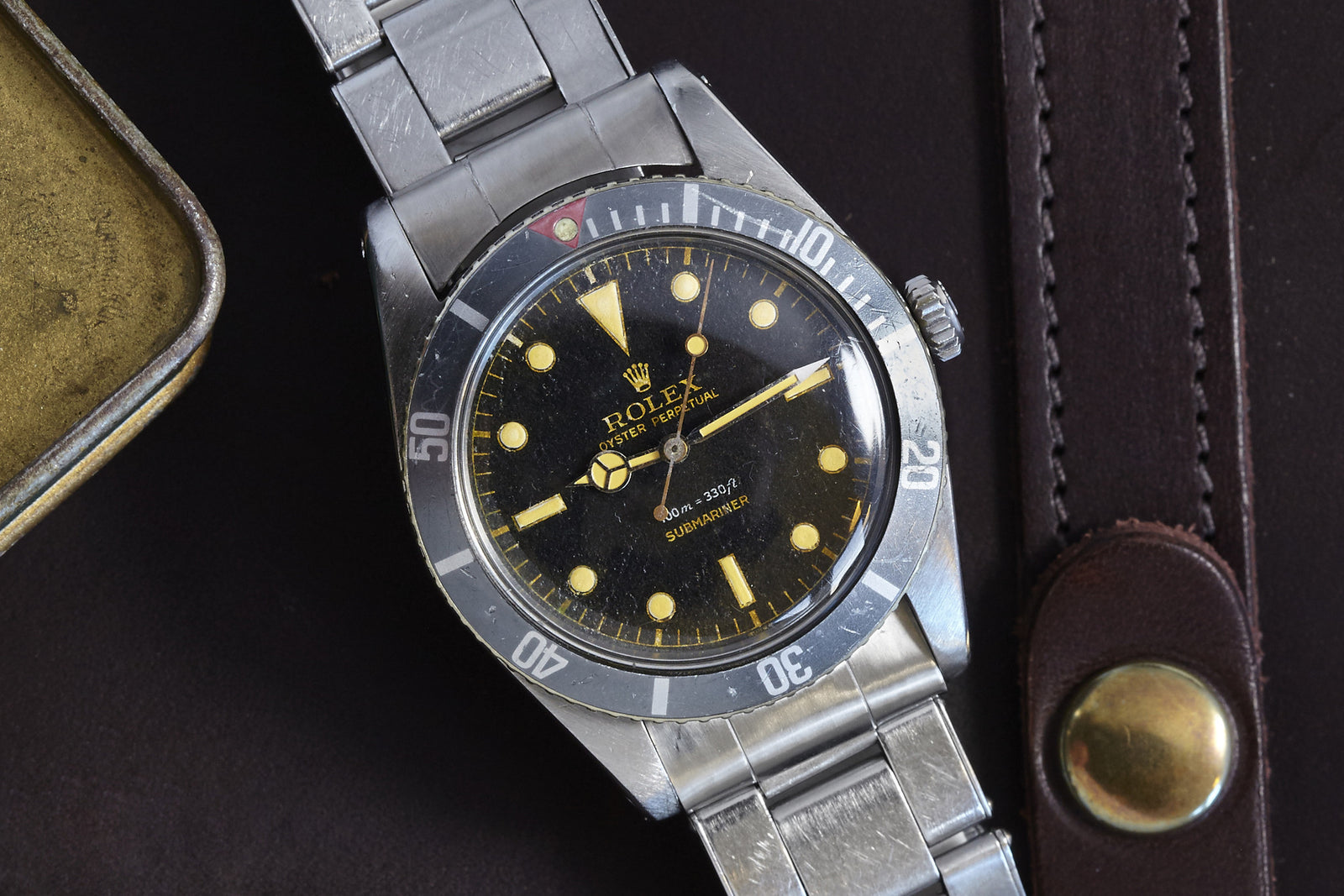 A close-up of the vintage Rolex Submariner Gilt Dial shows its black bezel, yellowed markers, and worn stainless steel band resting on dark leather, with a brown strap and brass case nearby.