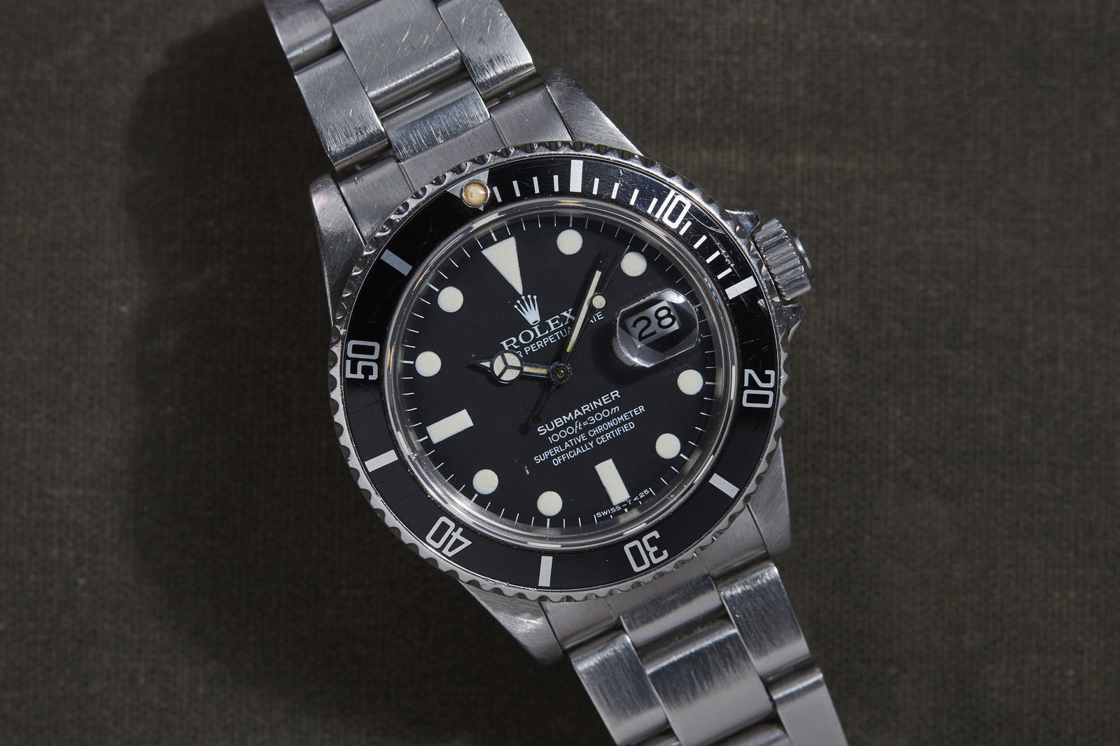 The Rolex Submariner by Rolex features a stainless steel case, black dial, white hour markers, and a date window at 3 o'clock, displayed on a dark textured surface.