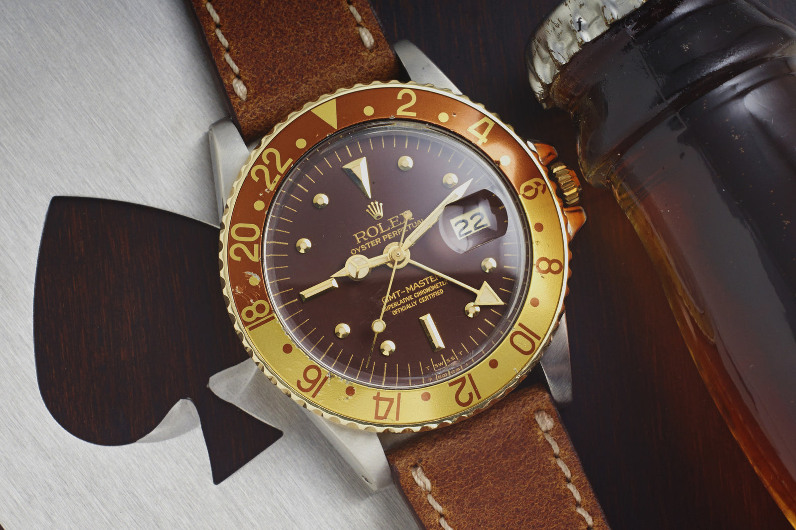 A Rolex Two-Tone "Root Beer" GMT Master from 1978, featuring a brown and gold bezel, brown dial, and leather strap, rests beside a dark bottle on a metallic surface with an axe-shaped cutout. The date on this steel timepiece shows 22.