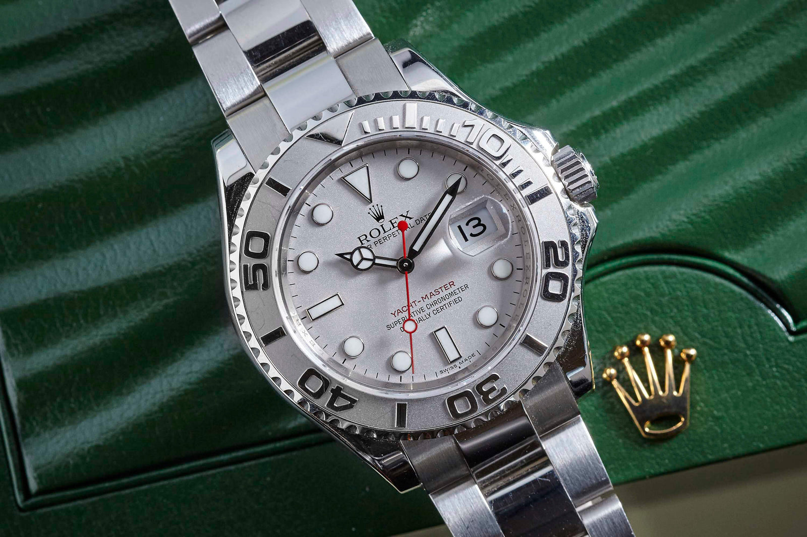 A close-up of the Rolex Yacht-Master by Rolex, featuring a white dial, bold hour markers, date window, and metal bracelet, displayed on a green box with the gold Rolex crown logo.