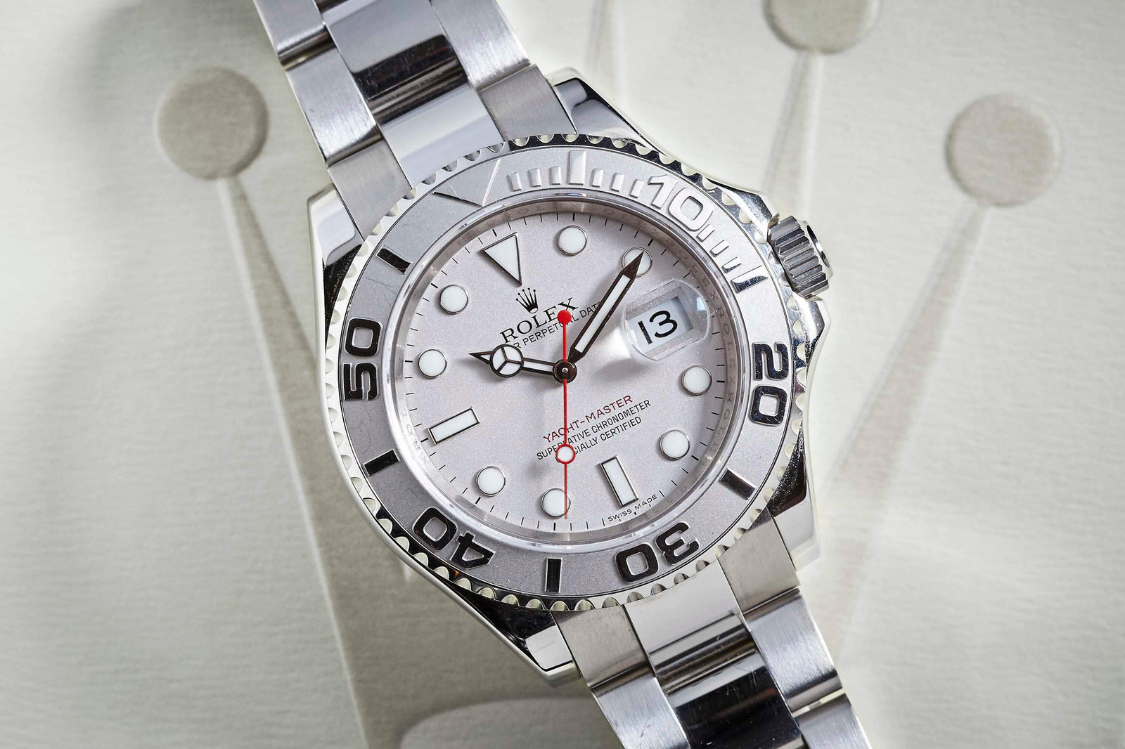 A close-up of the Rolex Yacht-Master by Rolex, featuring a silver case, white dial, black and silver bezel, red second hand, and metal bracelet resting on a textured surface with part of the Rolex crown logo visible.