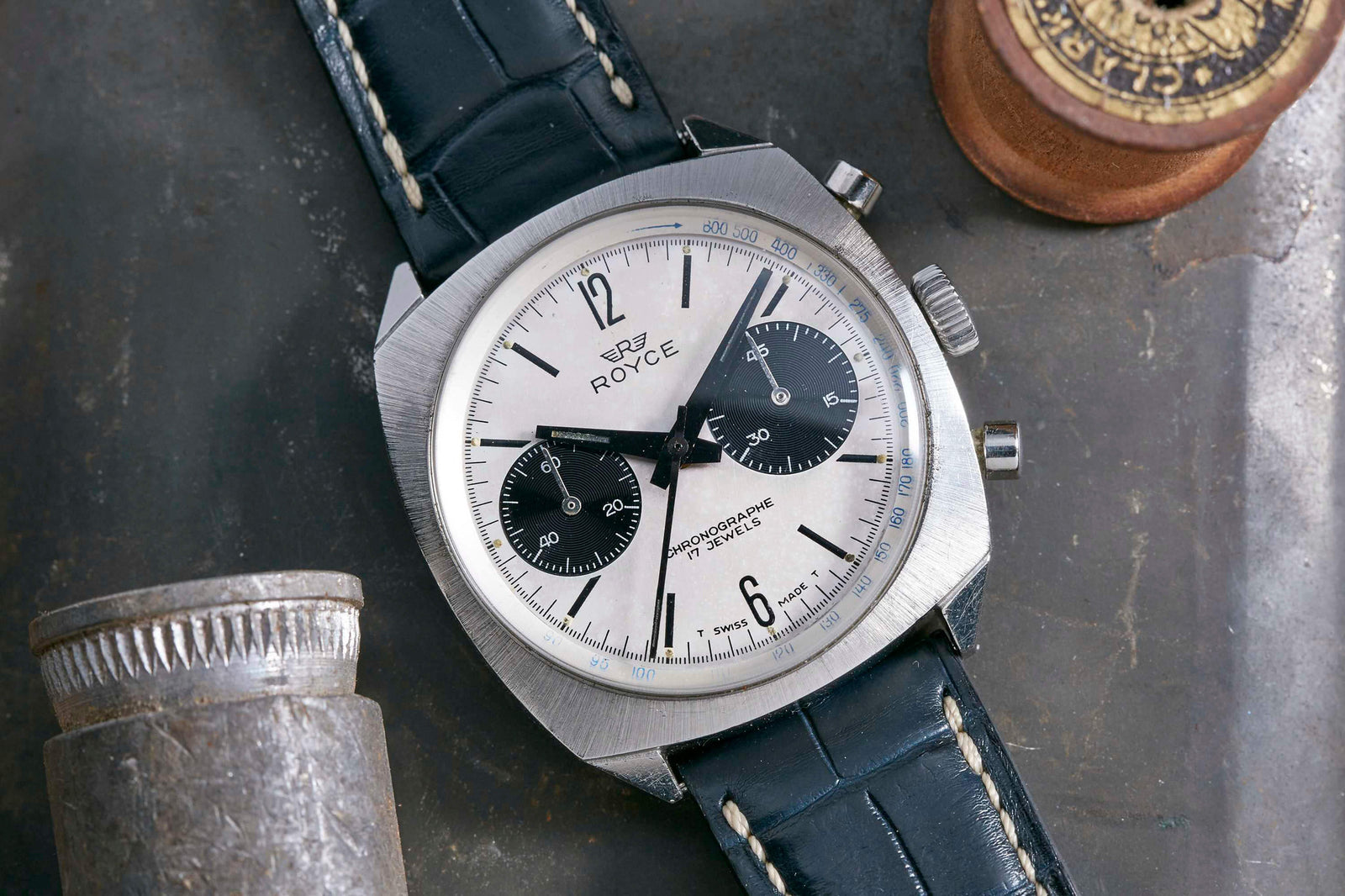 The Royce Panda Chronograph by Royce, a vintage wristwatch featuring a silver case, black leather strap, and two black subdials, is displayed on a rustic metal surface next to a metallic cylinder and round wooden object.