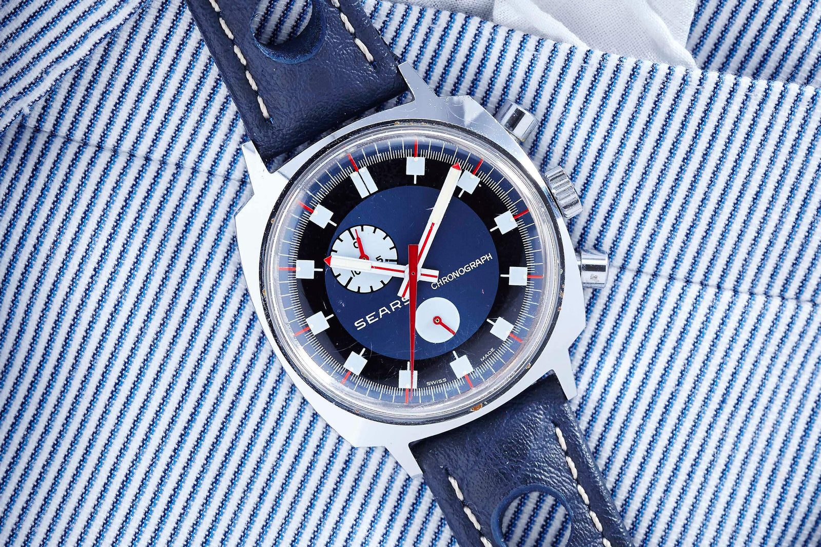 A close-up of a vintage 70s Sears Exotic Dial Chronograph with a blue leather strap resting on blue-and-white striped fabric. The watch has red, white, and blue accents, two subdials, and silver pushers.