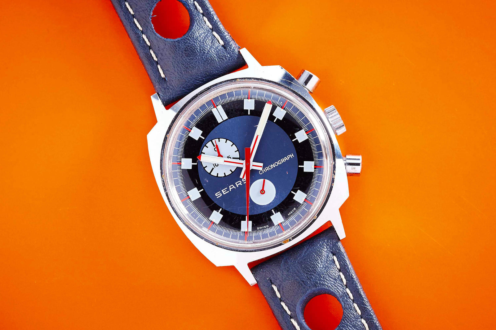 The Sears Exotic Dial Chronograph is a vintage 70s watch featuring a blue strap, black and white dial, red and white hands, two subdials, and an eye-catching vibrant orange background.