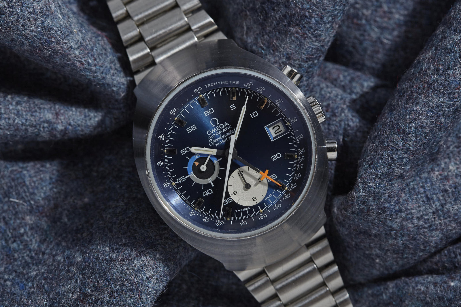A close-up of the Omega Speedmaster Mark III by Omega, featuring a blue dial, silver case, and metal bracelet on textured grey fabric; its face showcases multiple subdials and a date window—perfect for collectors.