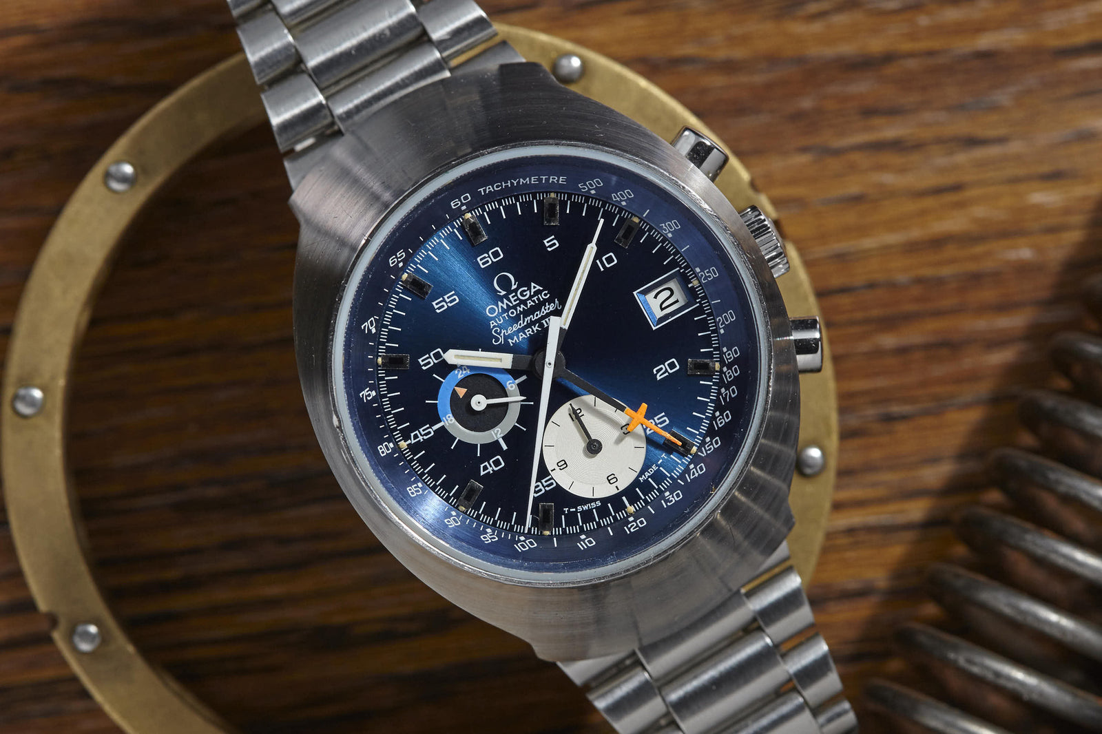 A close-up of an Omega Speedmaster Mark III by Omega features a blue dial, tachymeter bezel, date window, and silver stainless steel bracelet on a wooden surface with metal objects—making it a true collector’s watch.