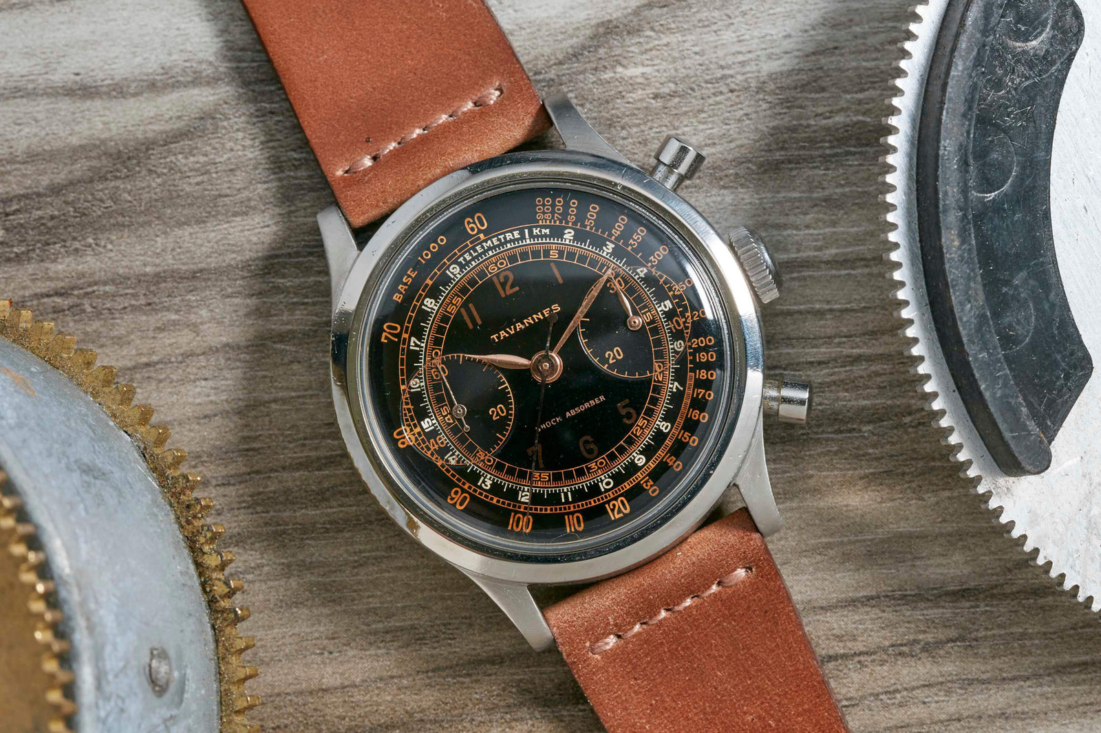 A close-up of a Tavannes Chronograph by Tavannes, featuring a brown leather strap, black dial, and gold numerals, elegantly placed on a wooden surface beside metallic gears.